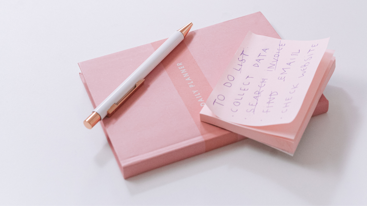 A pink planner with a white and rose gold pen resting on top, and a handwritten note on a curl of paper next to it that reads 'To do list: collect data, research email, fill out email website'.