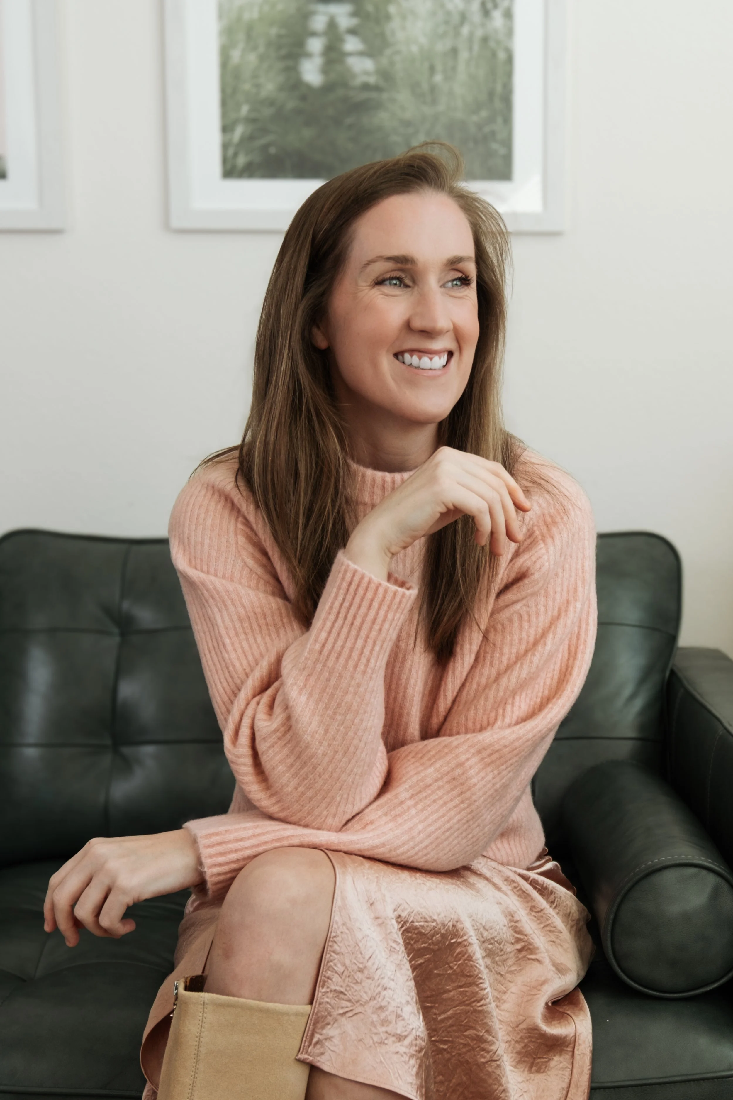 A woman with long brown hair smiling, sitting on a black leather couch in a pinkish beige sweater and skirt, in a room with white walls and framed pictures.