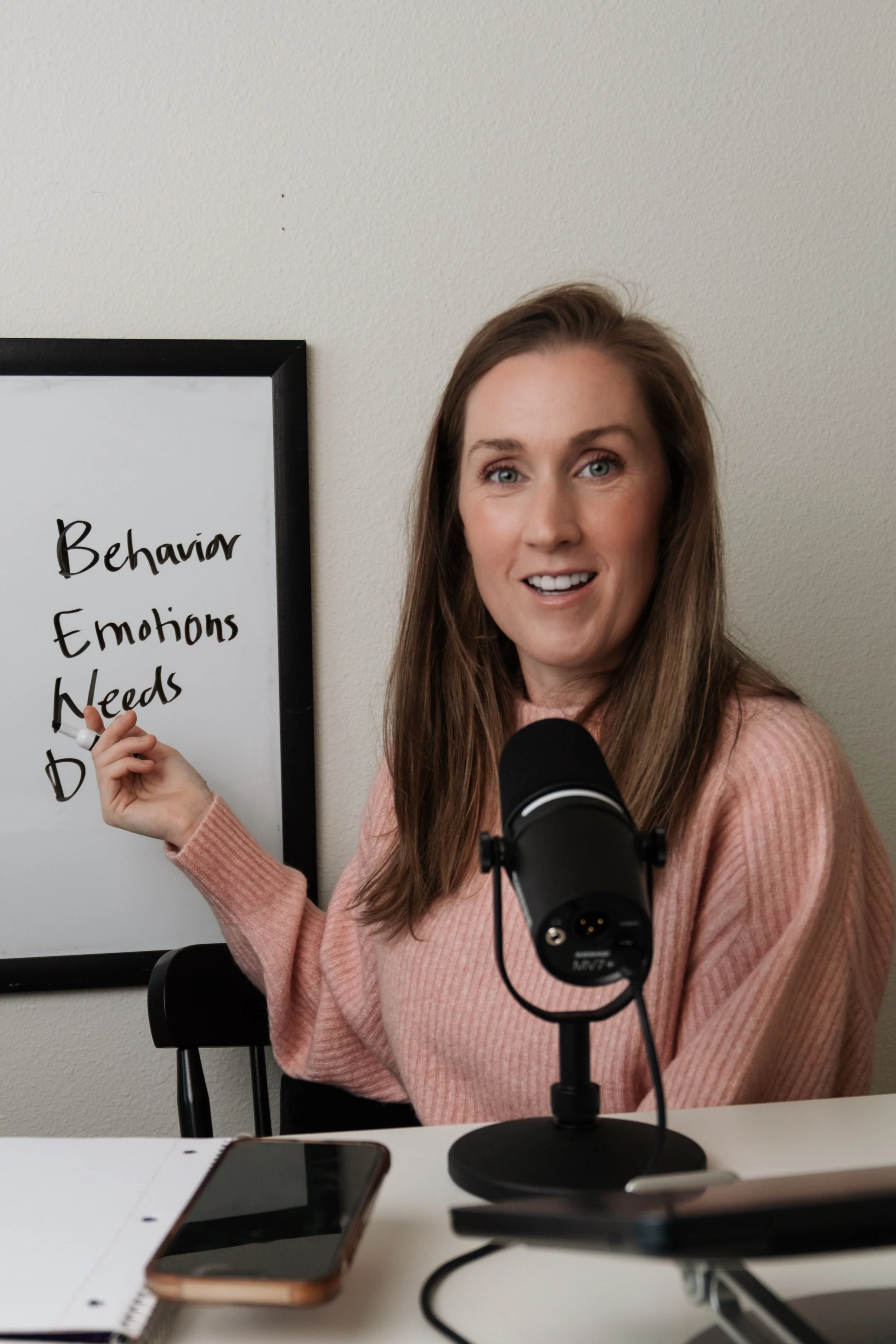 A woman with long brown hair in a pink sweater sitting at a desk in front of a microphone, pointing to a whiteboard with the words "Behavior, Emotions, Needs" written on it.