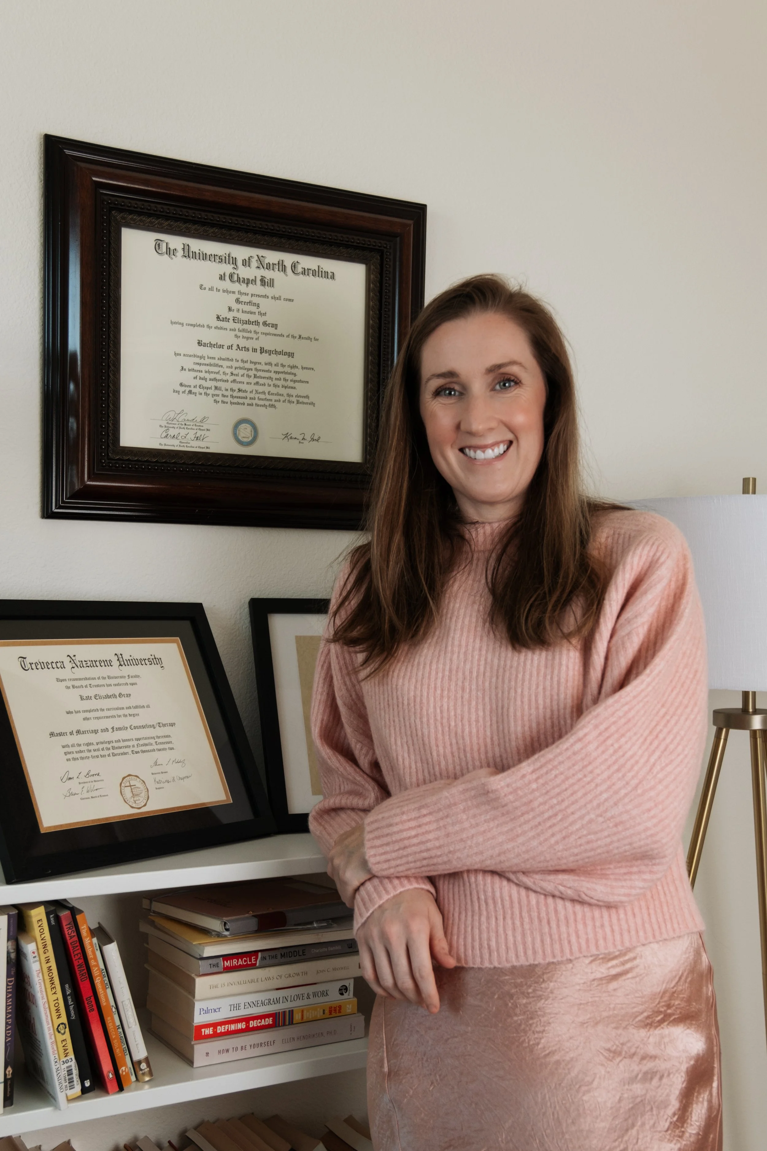 A woman with long brown hair smiling, standing in front of a wall with framed certificates and a bookshelf with books.
