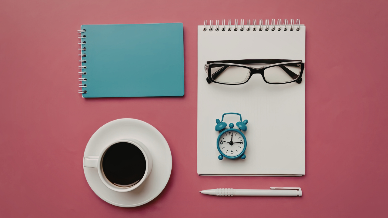 Flat lay image of a pink background with a blue notebook, white notepad with glasses and alarm clock on top, cup of coffee on saucer, and a white pen.