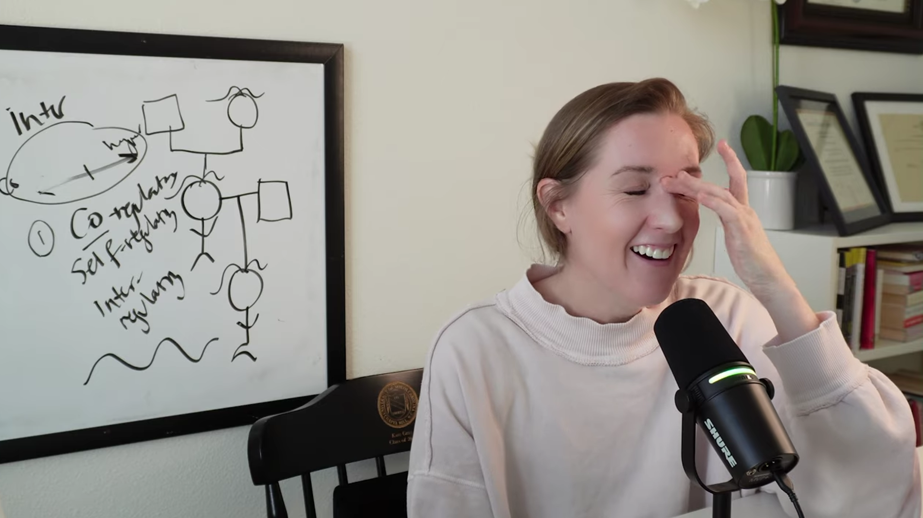 A woman with short brown hair is smiling and laughing, touching her forehead with her right hand while sitting at a desk with a microphone in front of her. Behind her, there is a whiteboard with handwritten notes and diagrams, and a bookshelf with framed documents and books.