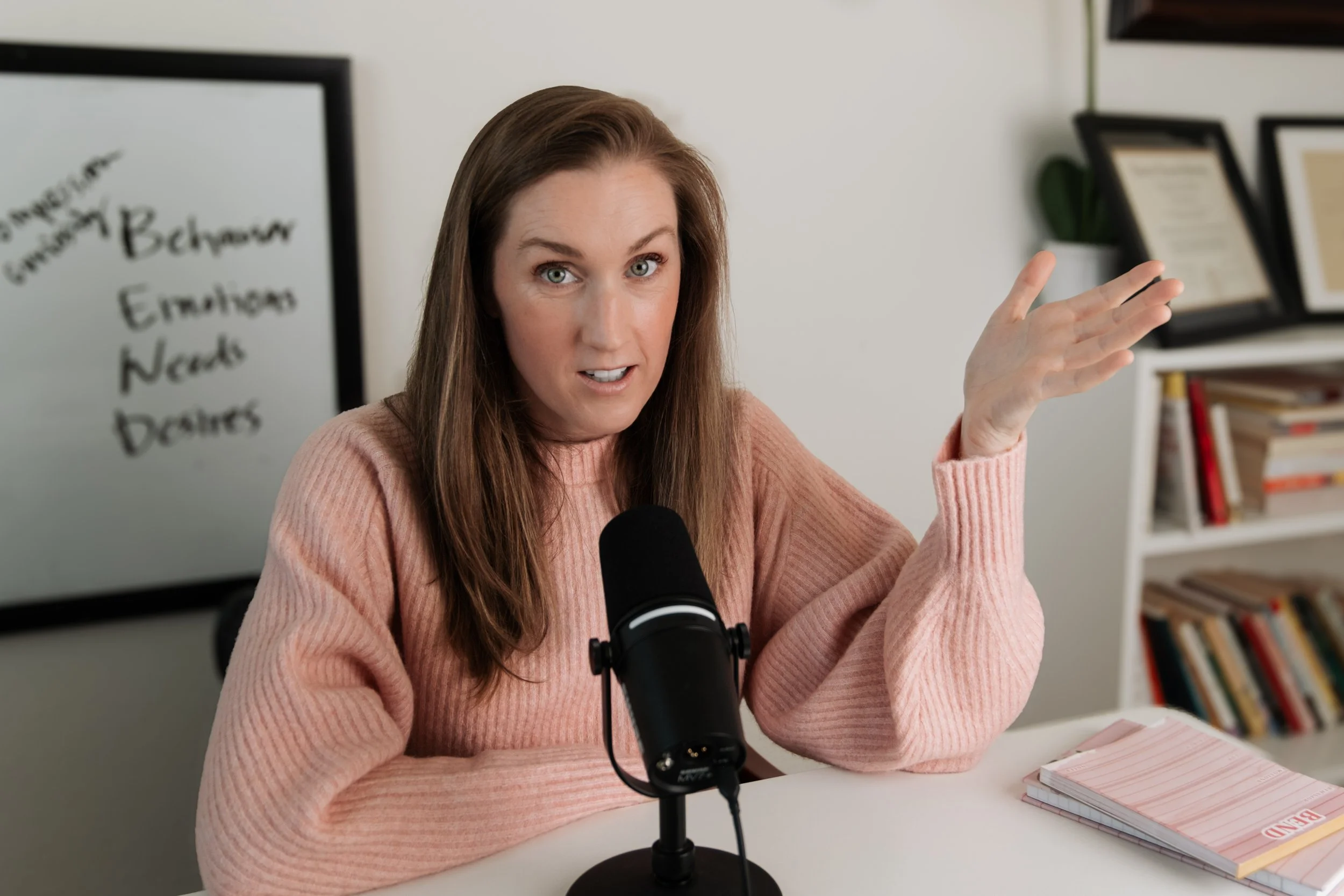 A woman with long brown hair wearing a pink sweater speaking into a microphone during a podcast recording or interview, with a whiteboard in the background listing words like 'Behavior,' 'Emotions,' 'Needs,' and 'Desires', surrounded by a bookshelf a
