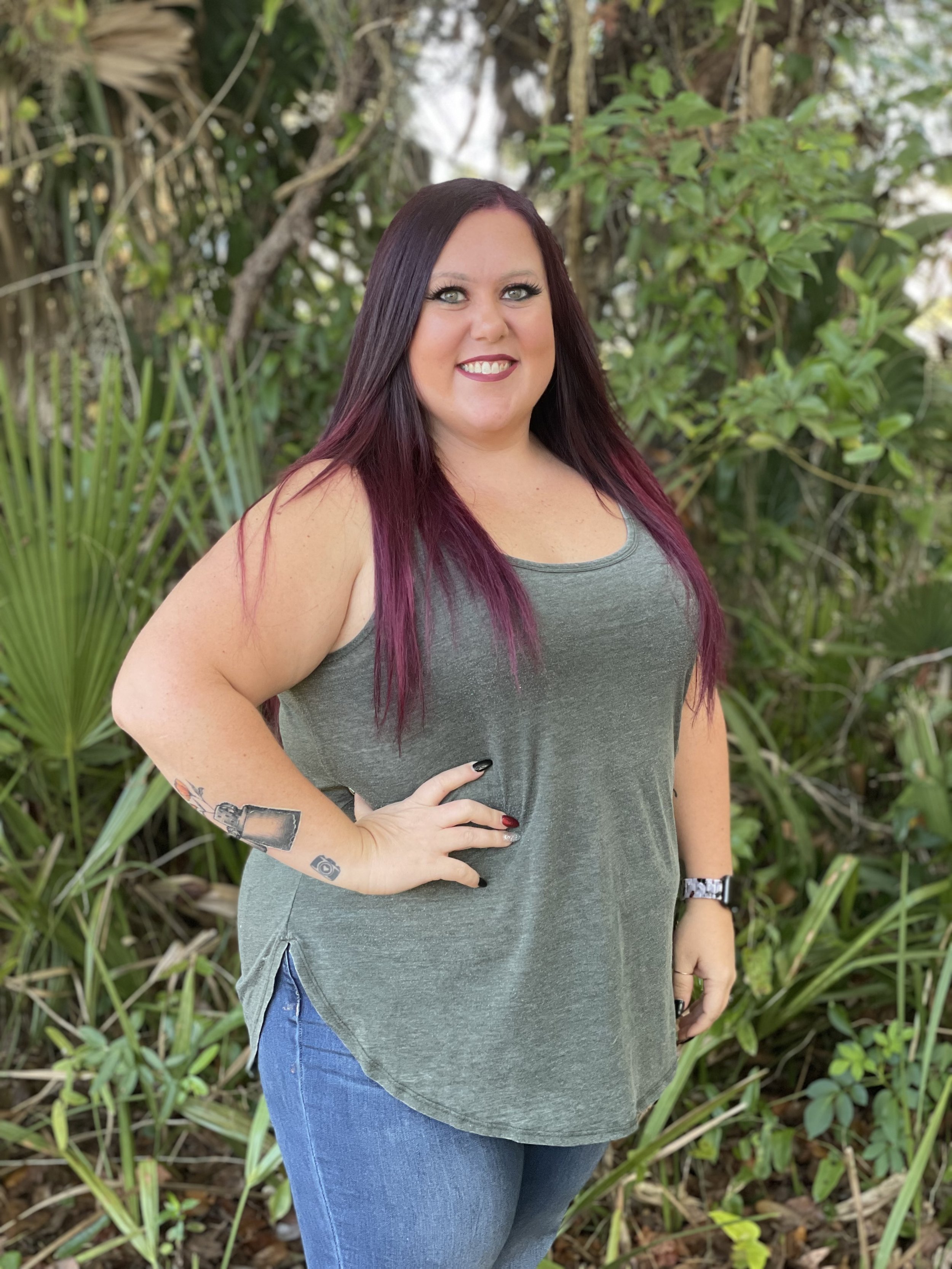 A cheerful woman with long, dark purple hair with pink tips, wearing a sleeveless gray top and blue jeans, standing outdoors in a lush, green setting with various plants and trees.