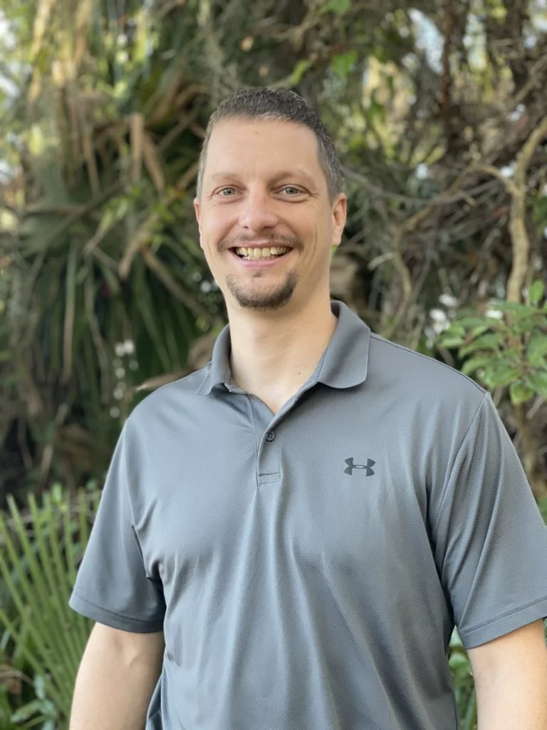 A smiling man with short dark hair and a goatee wearing a gray Under Armour polo shirt stands outdoors in front of green tropical plants.