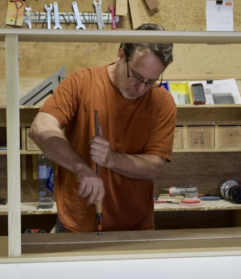 A man working on a woodworking project in a workshop, using a tool on a wooden piece, with various tools and supplies seen in the background.