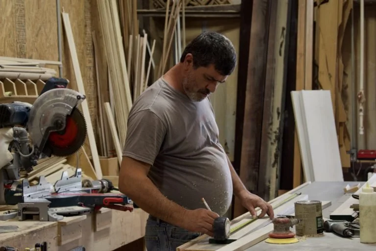 Man working on woodworking project in a workshop, surrounded by tools, wood pieces, and equipment.