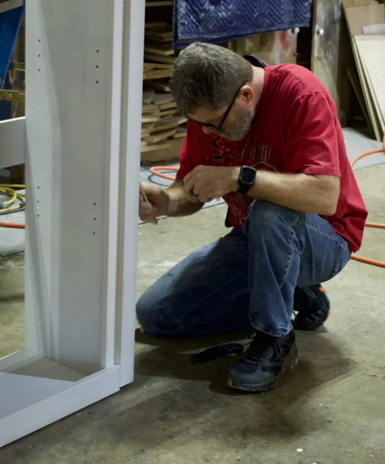 A man in a red shirt, glasses, and jeans kneeling on the workshop floor, working on assembling or repairing a piece of furniture or equipment.