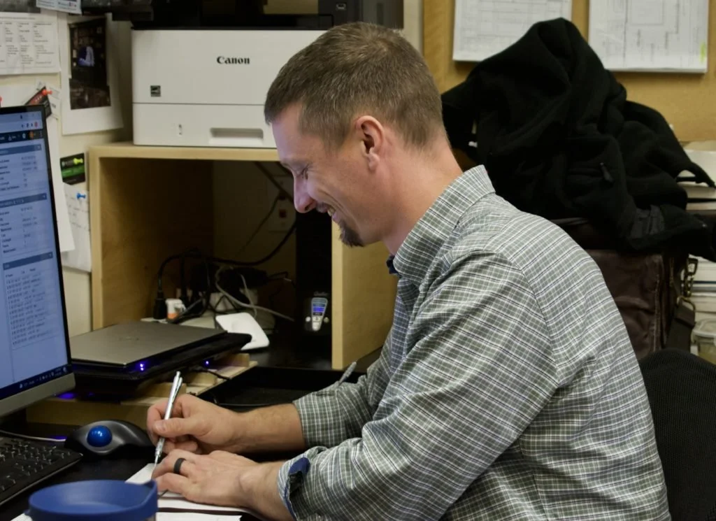 A man sitting at a desk, smiling, taking notes, with computer equipment and a printer in an office setting.