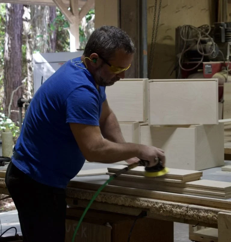 Man in blue shirt and yellow safety glasses sanding wooden pieces in a workshop with woodworking tools and stacks of wood.