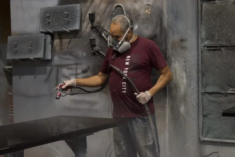 A man wearing a mask and gloves spray-paints a surface in a spray booth, with gray and black walls and equipment visible behind him.