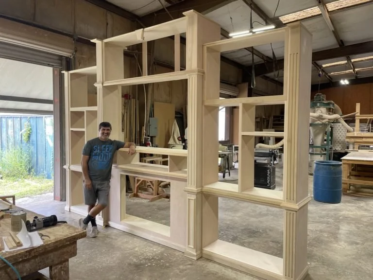 A man is standing and leaning against a large, partially assembled wooden bookshelf or room divider in a woodworking workshop. The structure has multiple open shelves and decorative columns. The workshop has various woodworking tools, materials, and other furniture projects in the background.