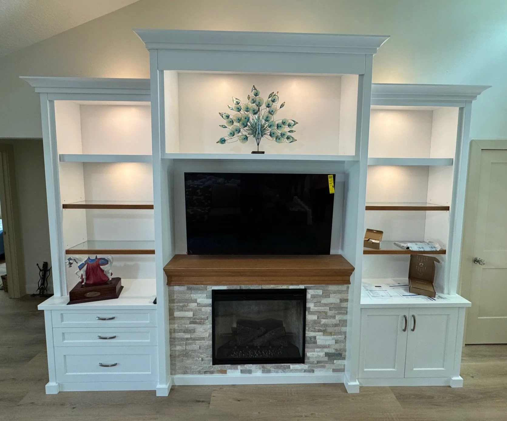 White entertainment center with built-in shelves, a mounted flat-screen TV, and a fireplace with stacked stone and a wooden mantel