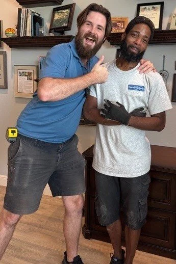 Two men standing in a room with framed pictures on the wall. One man, with light skin and a beard, is smiling and giving a thumbs up while placing his arm around the other man. The other man, with darker skin and wearing a work uniform and gloves, is holding a small item and smiling. The scene appears friendly and casual.