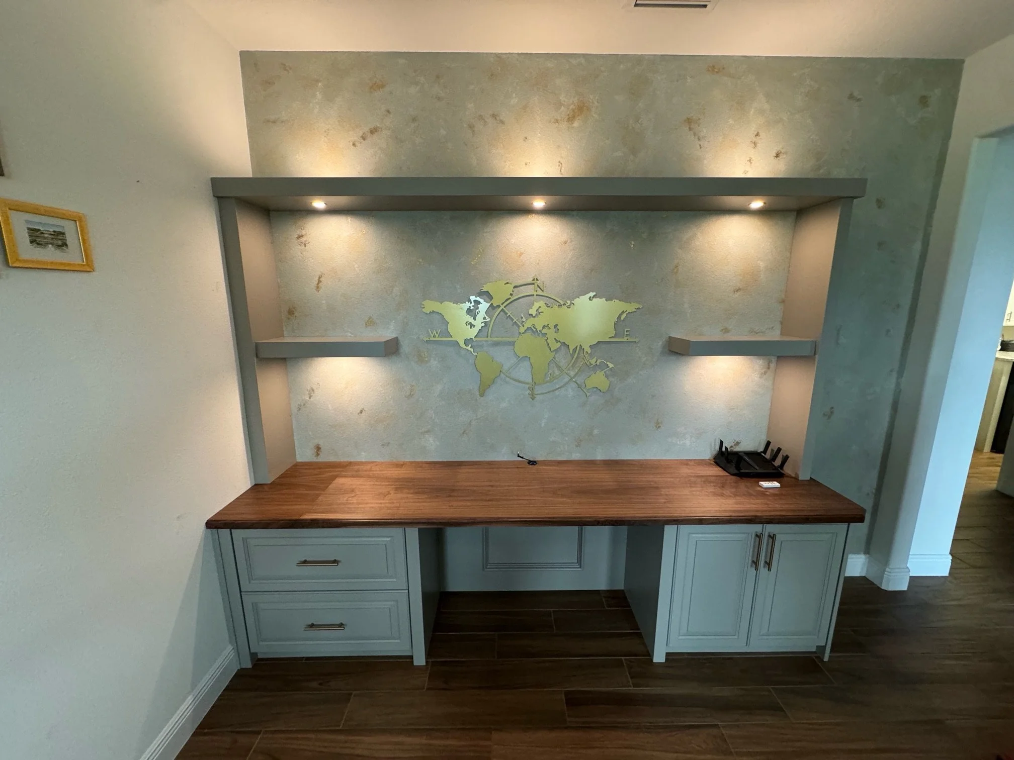A built-in desk with a wooden surface and light blue cabinets below, set against a textured wall with a world map decal and three small bookshelves. There are three ceiling lights illuminating the wall.