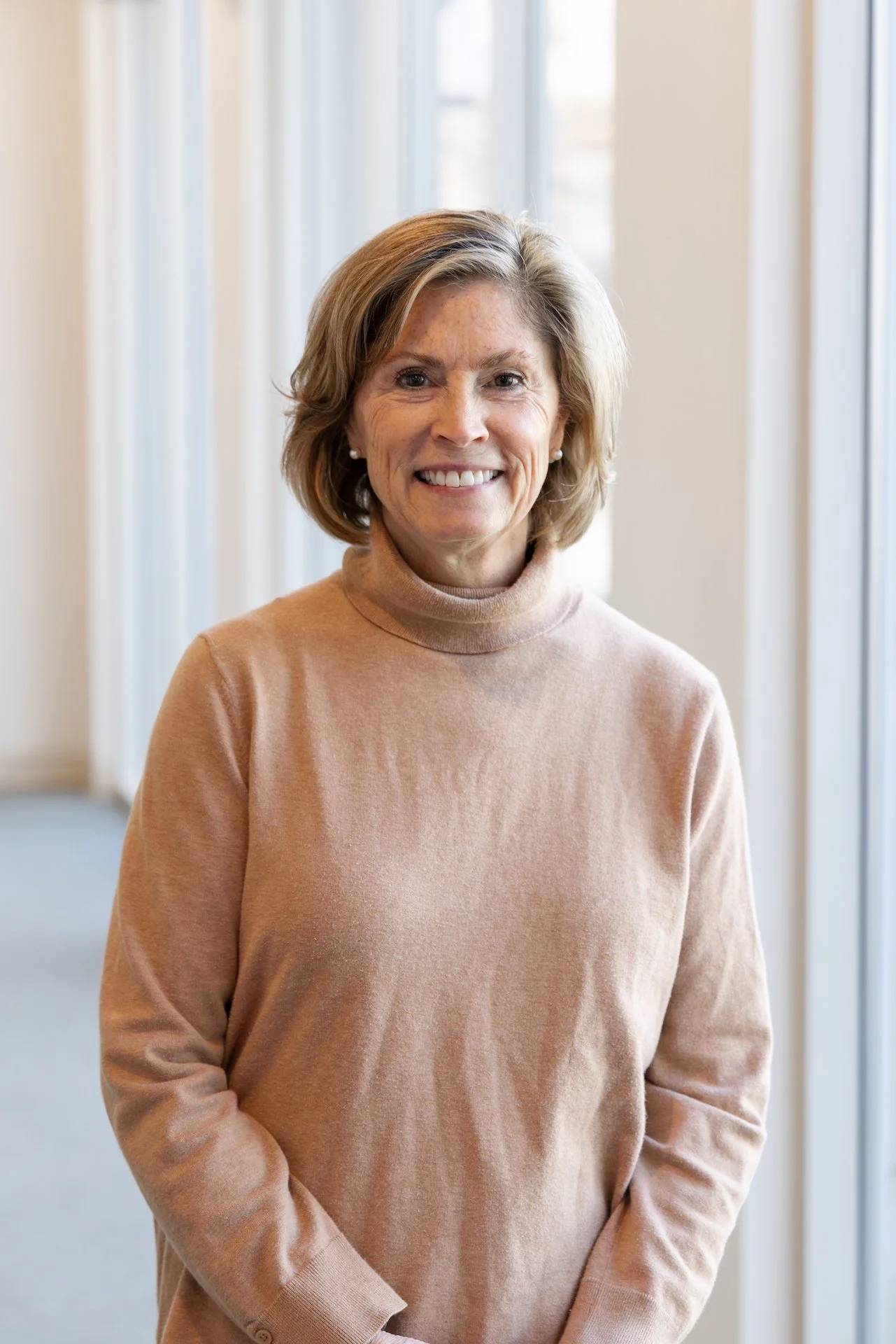 A middle-aged woman with short, wavy hair smiling while standing indoors by a large window.