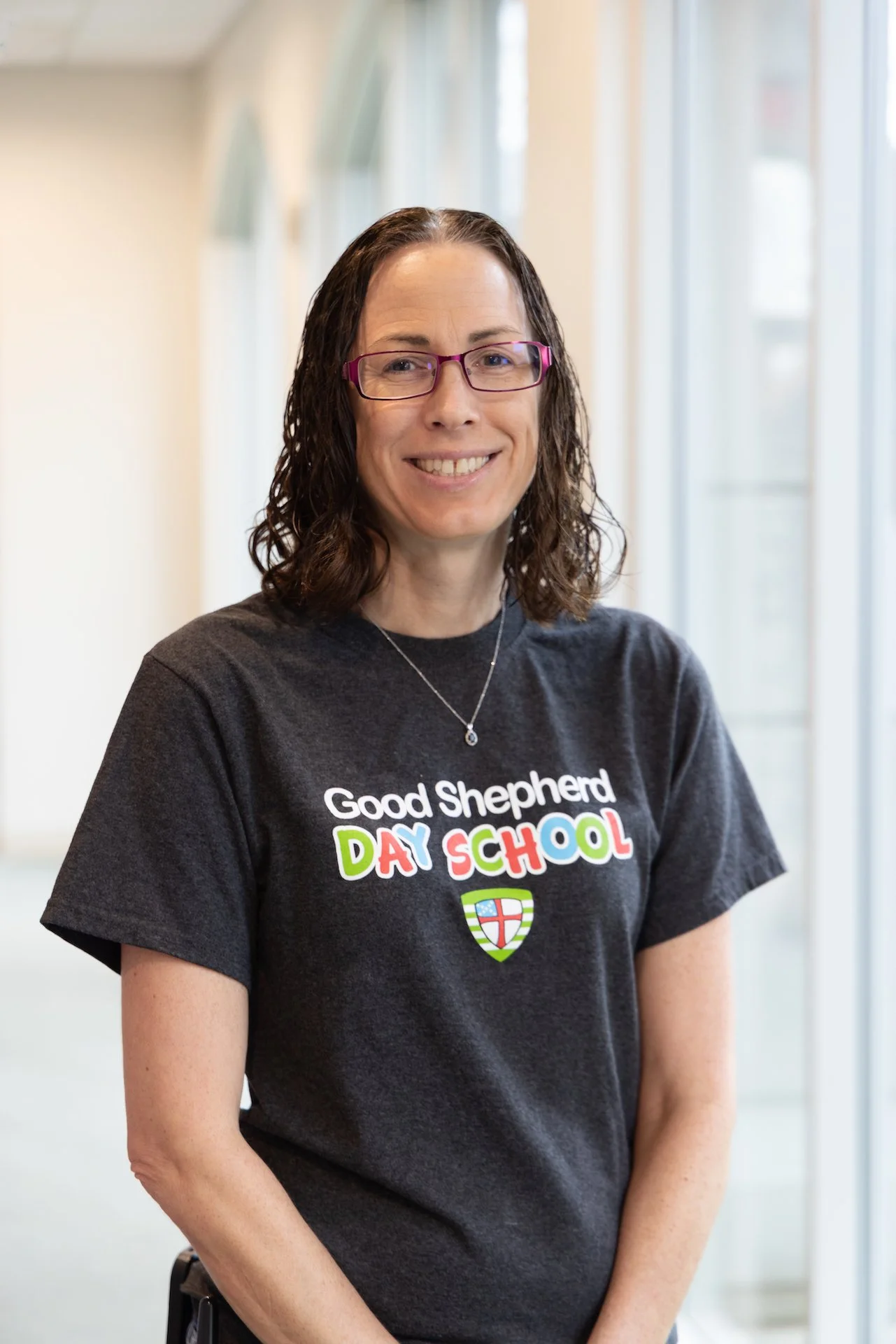 A woman with brown curly hair, wearing glasses and a dark gray T-shirt that says 'Good Shepherd Day School' with colorful letters and a shield emblem, standing in a corridor with large windows.