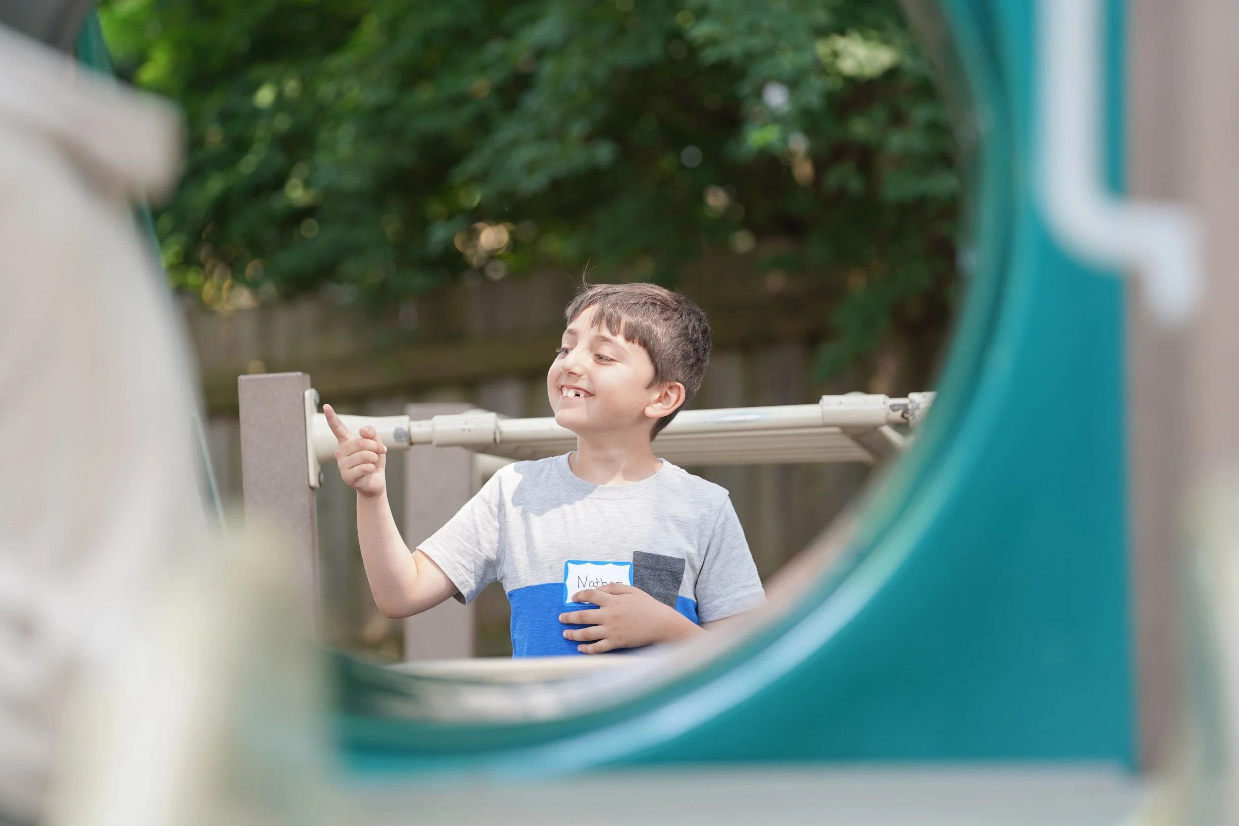 A young boy smiling and pointing upward while standing outdoors in front of a wooden fence and green trees, viewed through a playground tunnel.