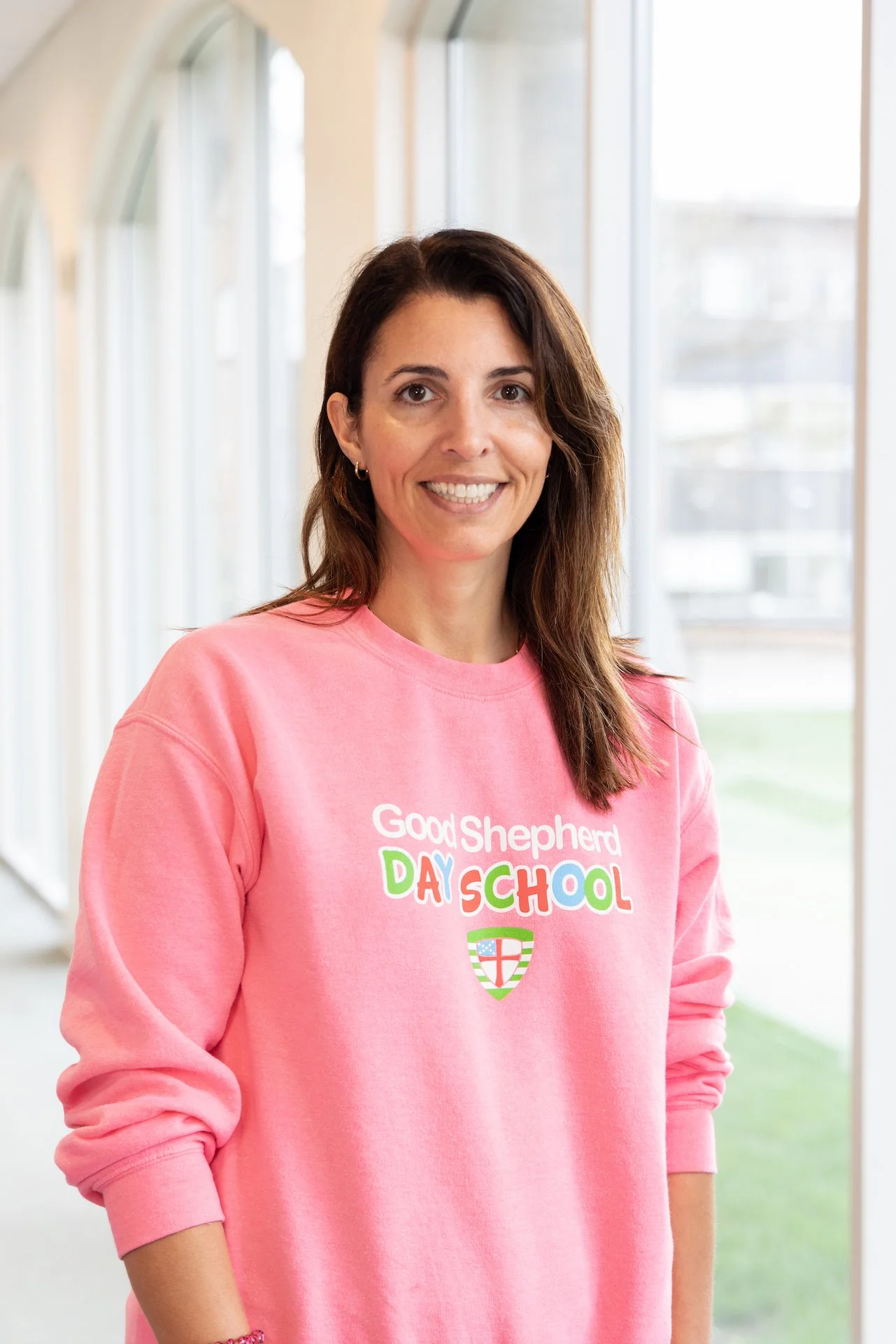 A woman with shoulder-length brown hair, wearing a pink sweatshirt that reads 'Good Shepherd Day School' with a crest, stands in front of large windows in a brightly lit room.