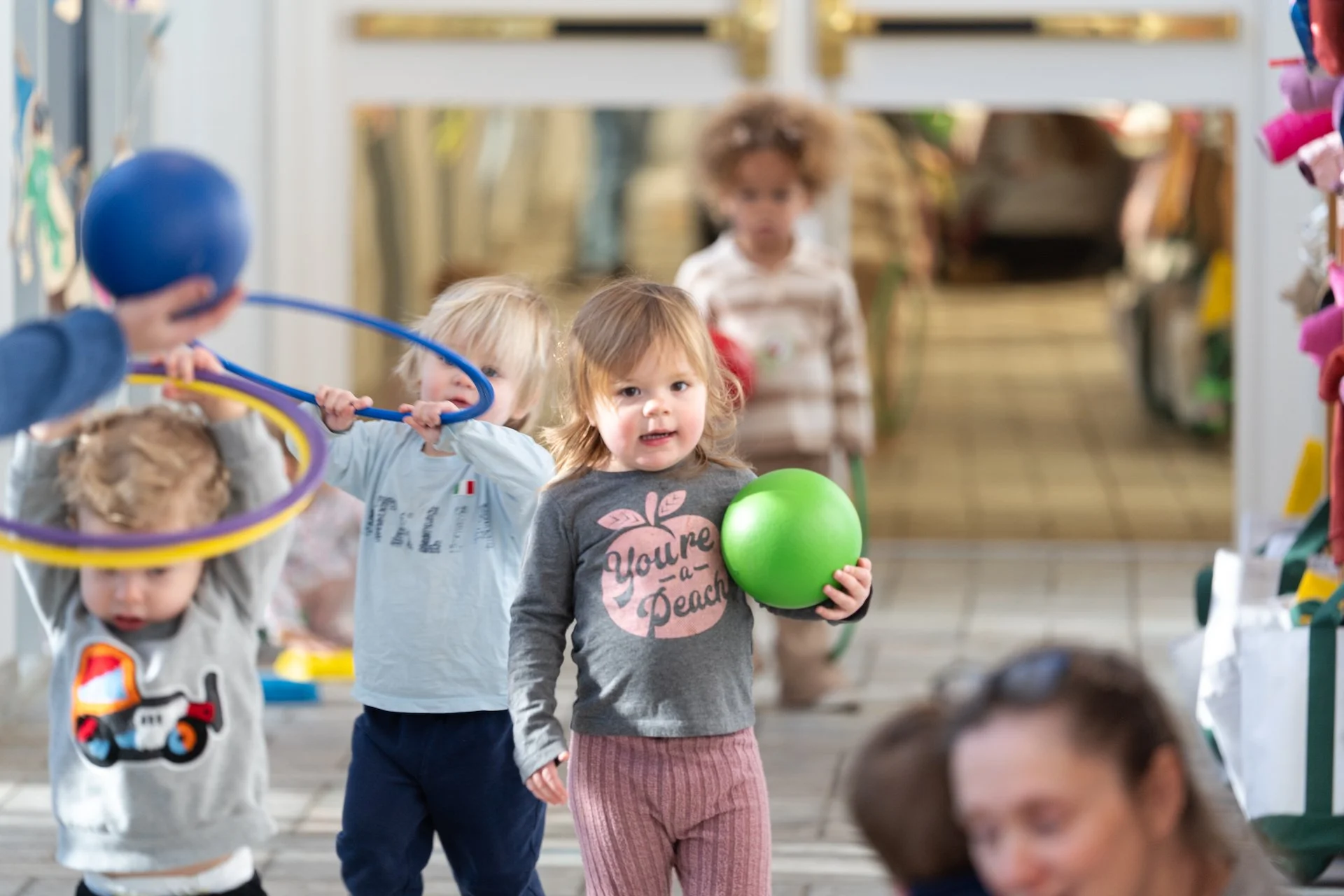 Children playing indoors, with one girl holding a green ball, while others play with hula hoops and observe.