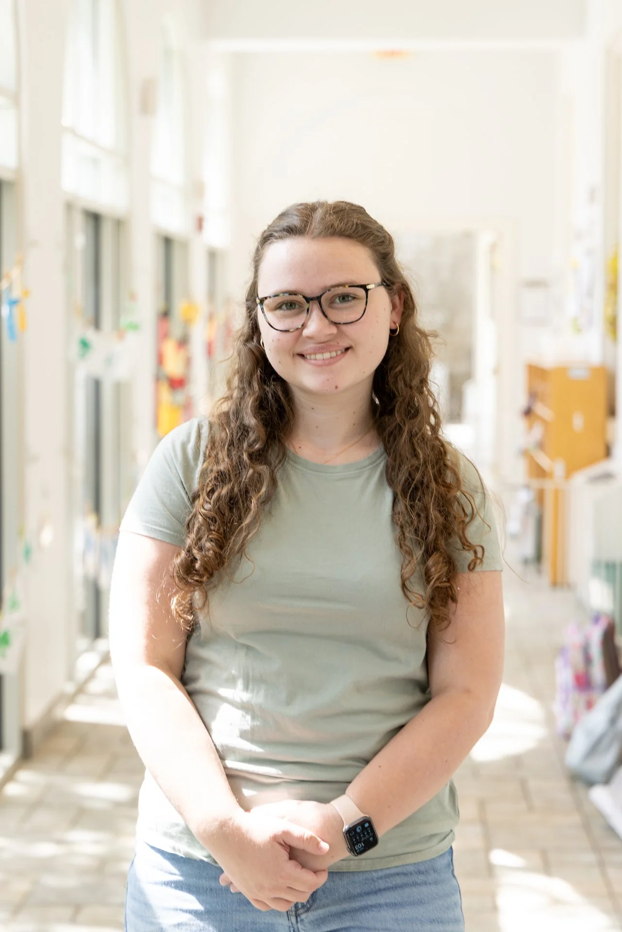 A young woman with long curly brown hair, glasses, and a light green t-shirt smiling in a bright hallway.