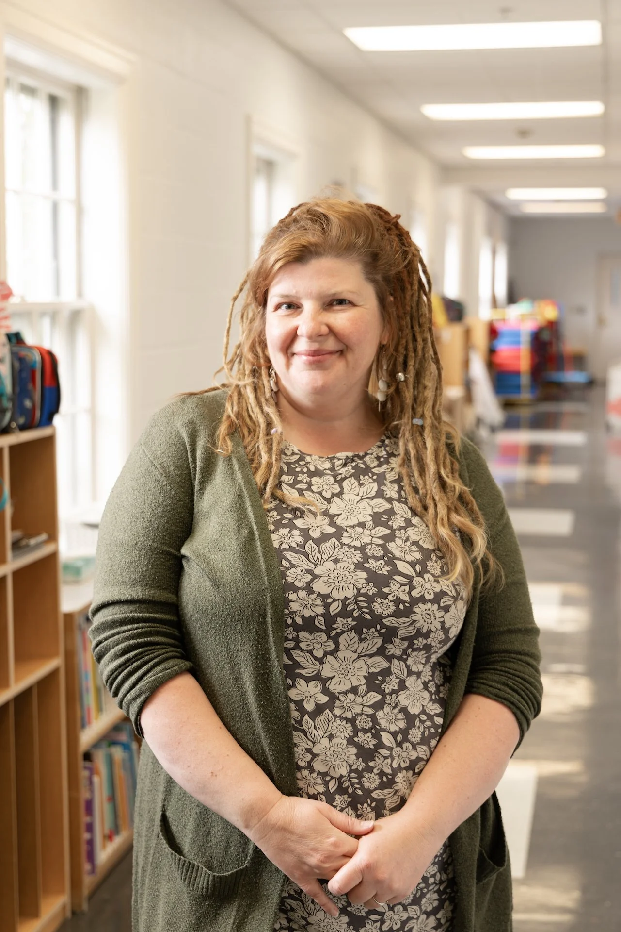 A woman with long, curly, reddish-brown hair and earrings standing in a well-lit school hallway filled with shelves of books and colorful backpacks in the background.
