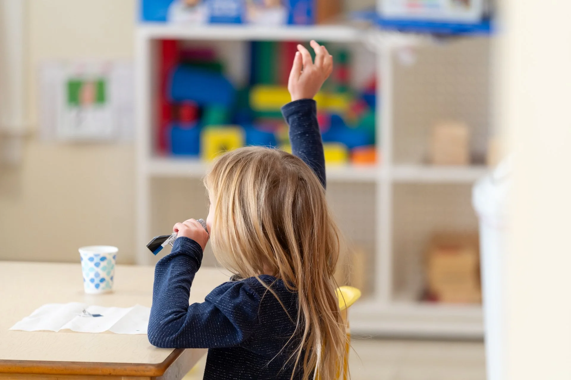 A young girl with long blond hair sitting at a table, raising her right hand, and holding a microphone in her left hand. There is a cup and some paper on the table, with a colorful bookshelf in the background.