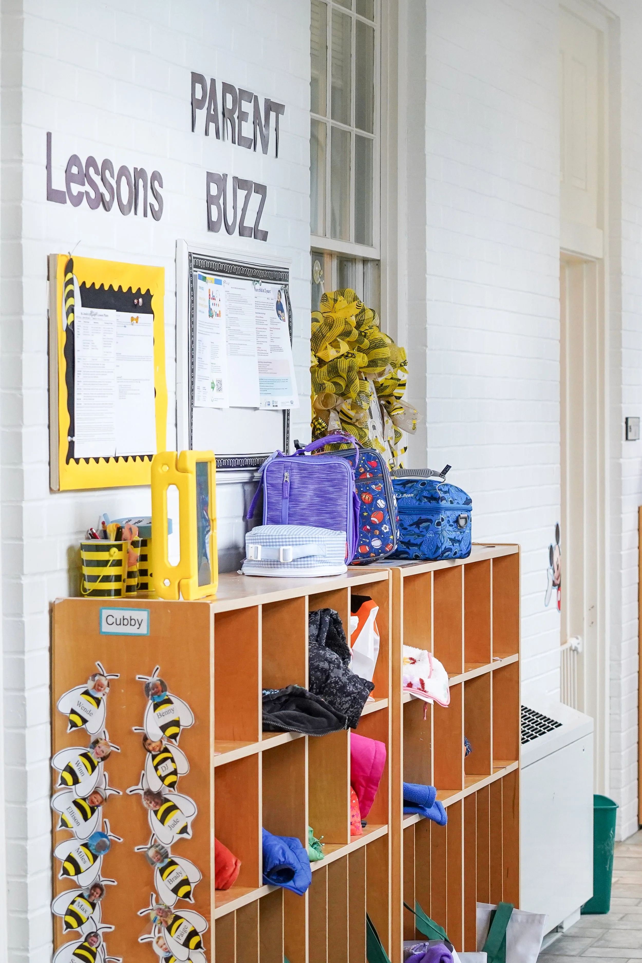 A classroom display with the sign 'Parent Lessons Buzz' on a white brick wall. There are cubbies with school bags and clothing, some bee-themed decorations, and papers posted on a bulletin board.