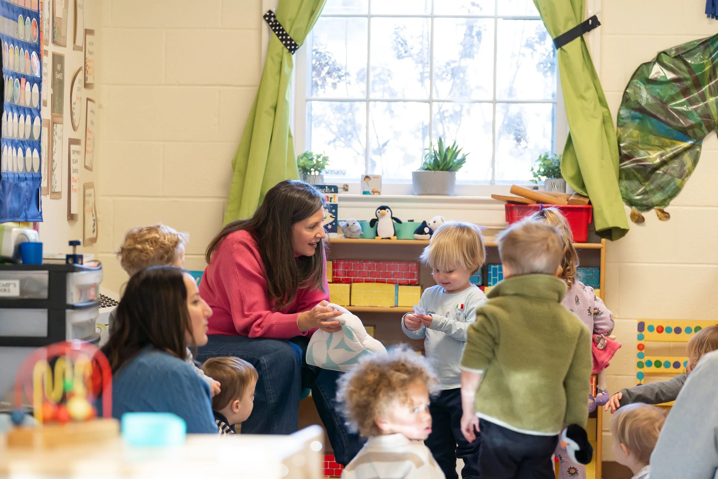 A preschool classroom with children and a teacher sitting and interacting near a window with green curtains. The room has colorful toys and decorations.