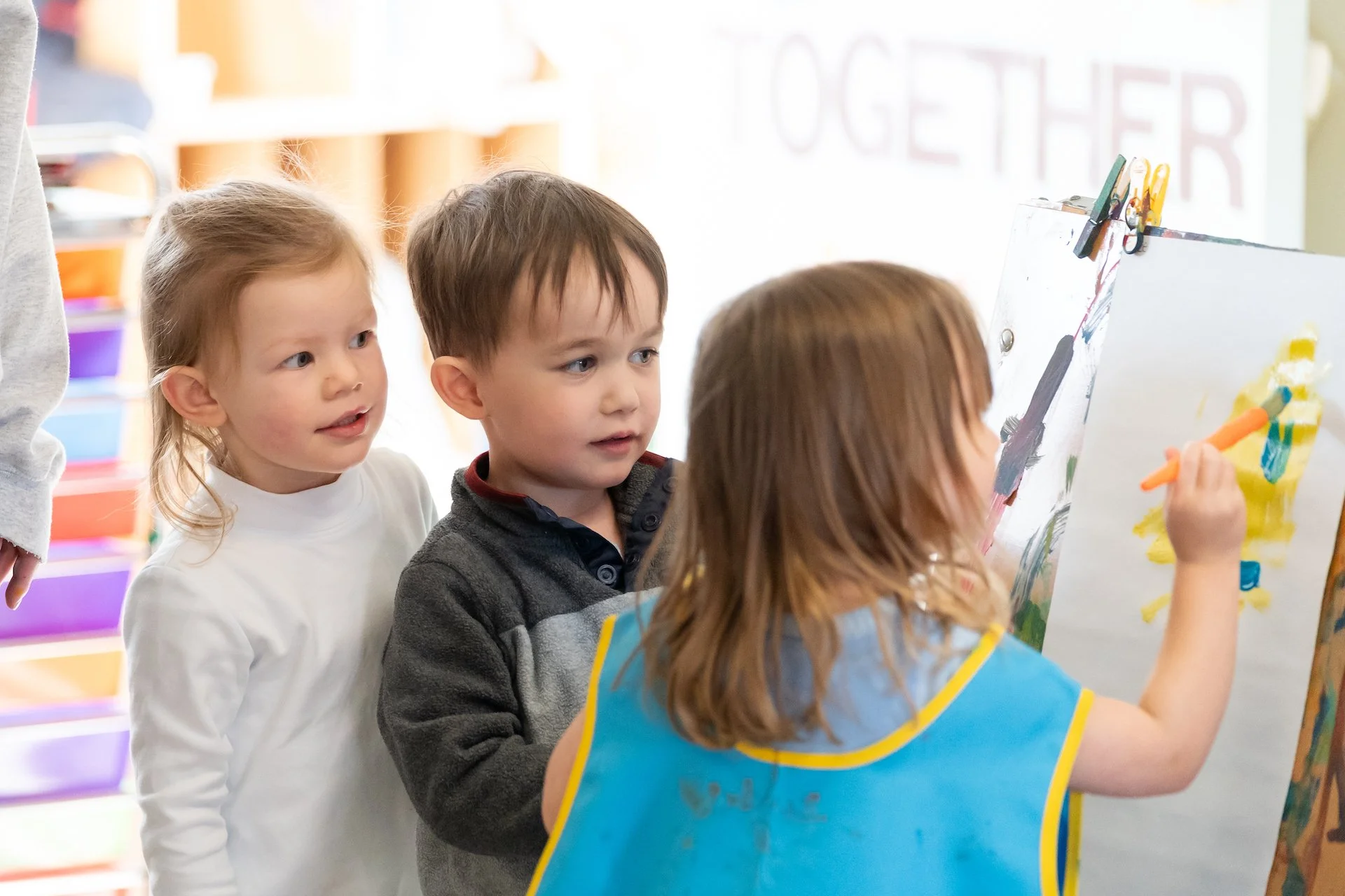 Three children standing in front of an easel, one girl painting with orange and yellow colors, two kids watching her. A colorful classroom backdrop is visible.
