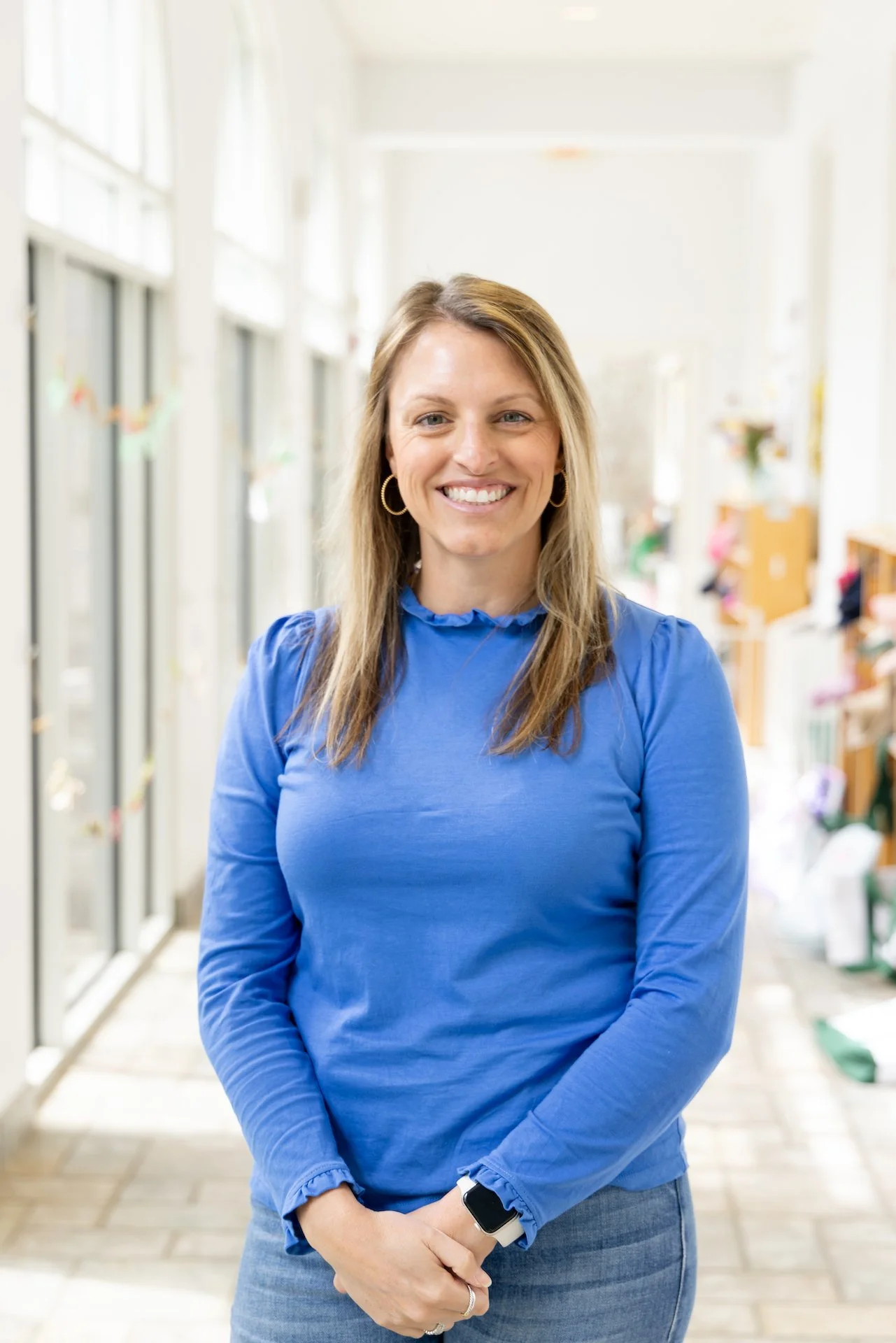 A smiling woman in a blue blouse standing in a brightly lit hallway.