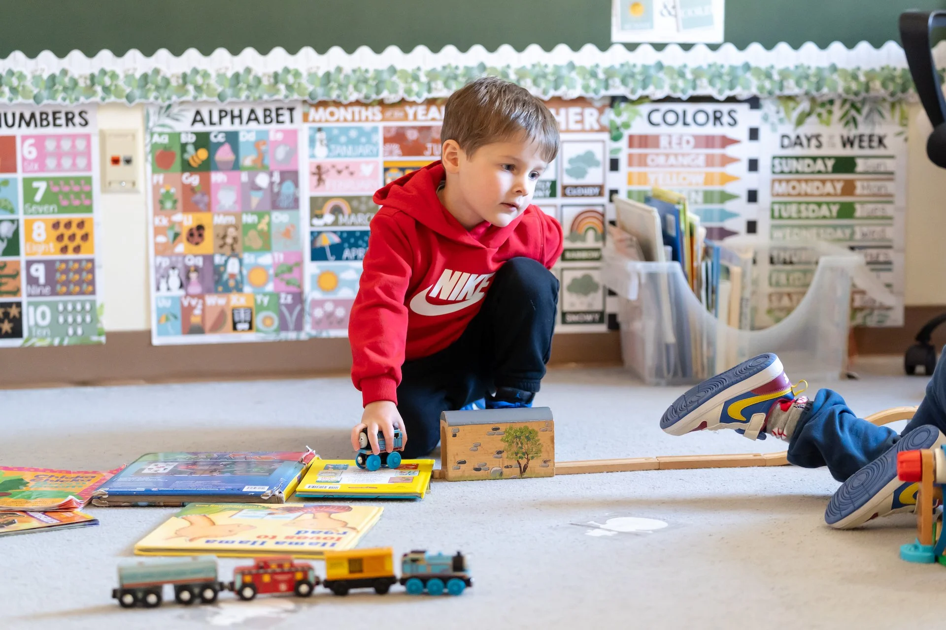 A young boy in a red Nike hoodie crouches on the floor playing with toy train tracks and a train. There are books and toys around him, with educational posters on the wall behind showing numbers, alphabet, months, weather, colors, and days of the week.