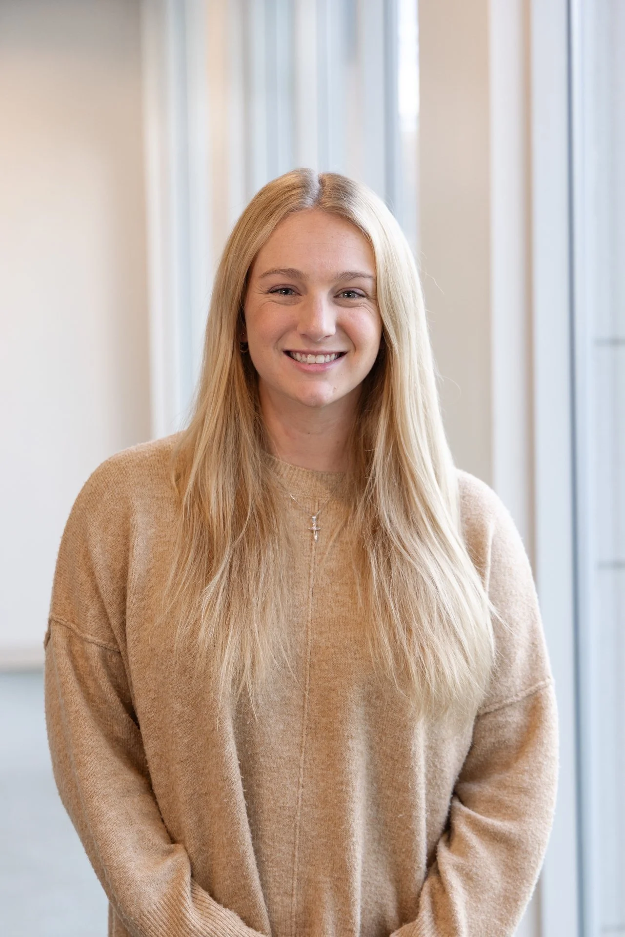 A young woman with long blonde hair, smiling, wearing a beige sweater and a necklace with a cross, standing indoors near large windows.