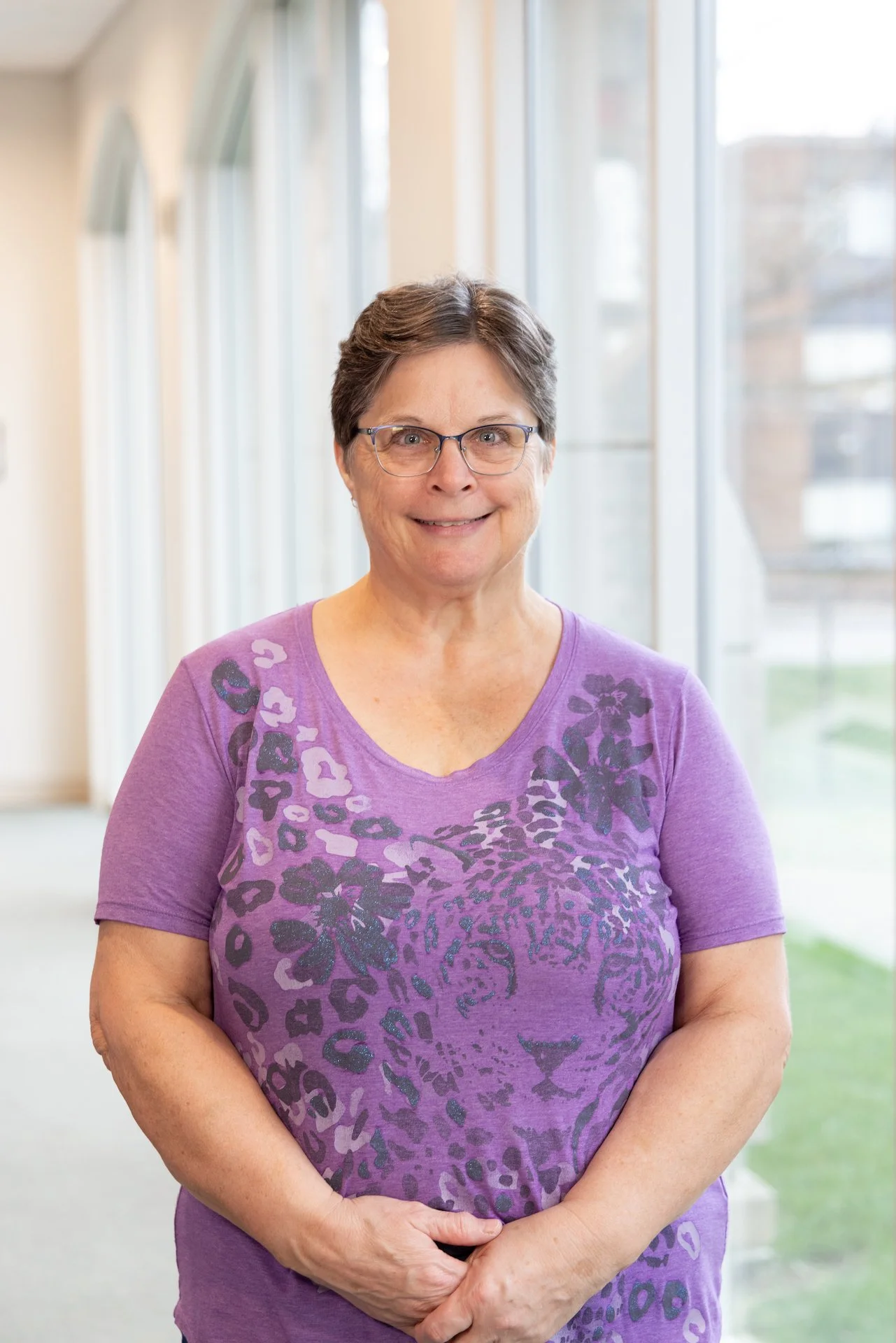 An older woman with short brown hair, glasses, and a purple leopard-print shirt standing in a brightly lit indoor space with large windows.
