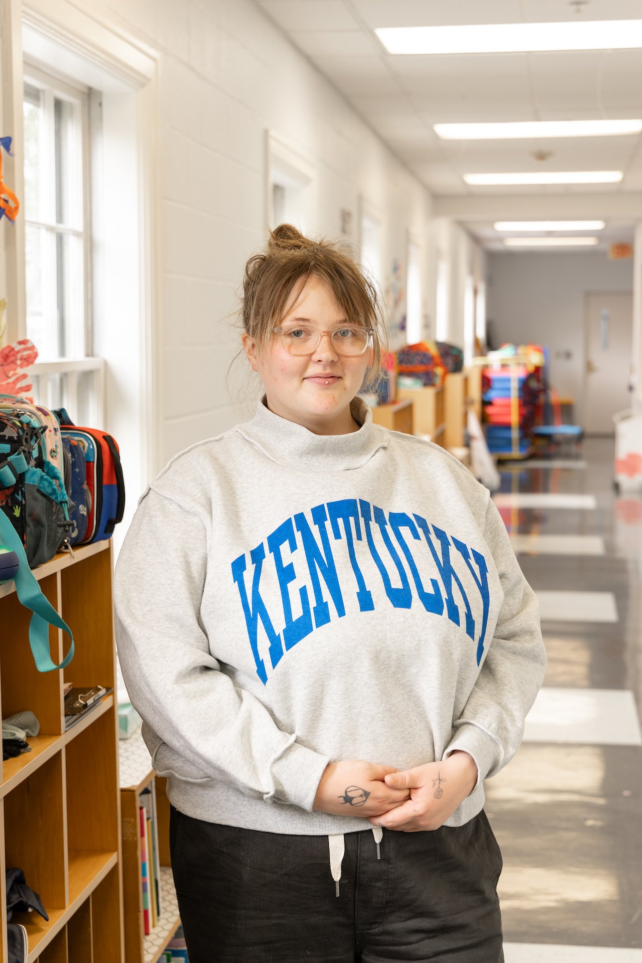 A young woman with glasses wearing a gray 'Kentucky' sweatshirt stands in a hallway with shelves of backpacks and school supplies.
