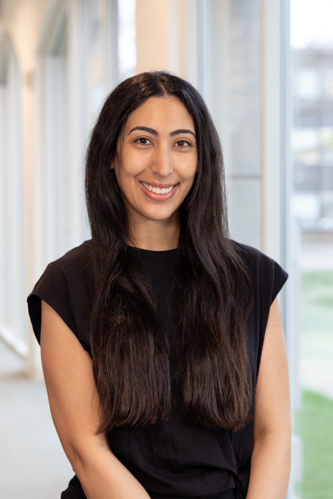Smiling woman with long dark hair wearing a black top standing indoors near a large window.