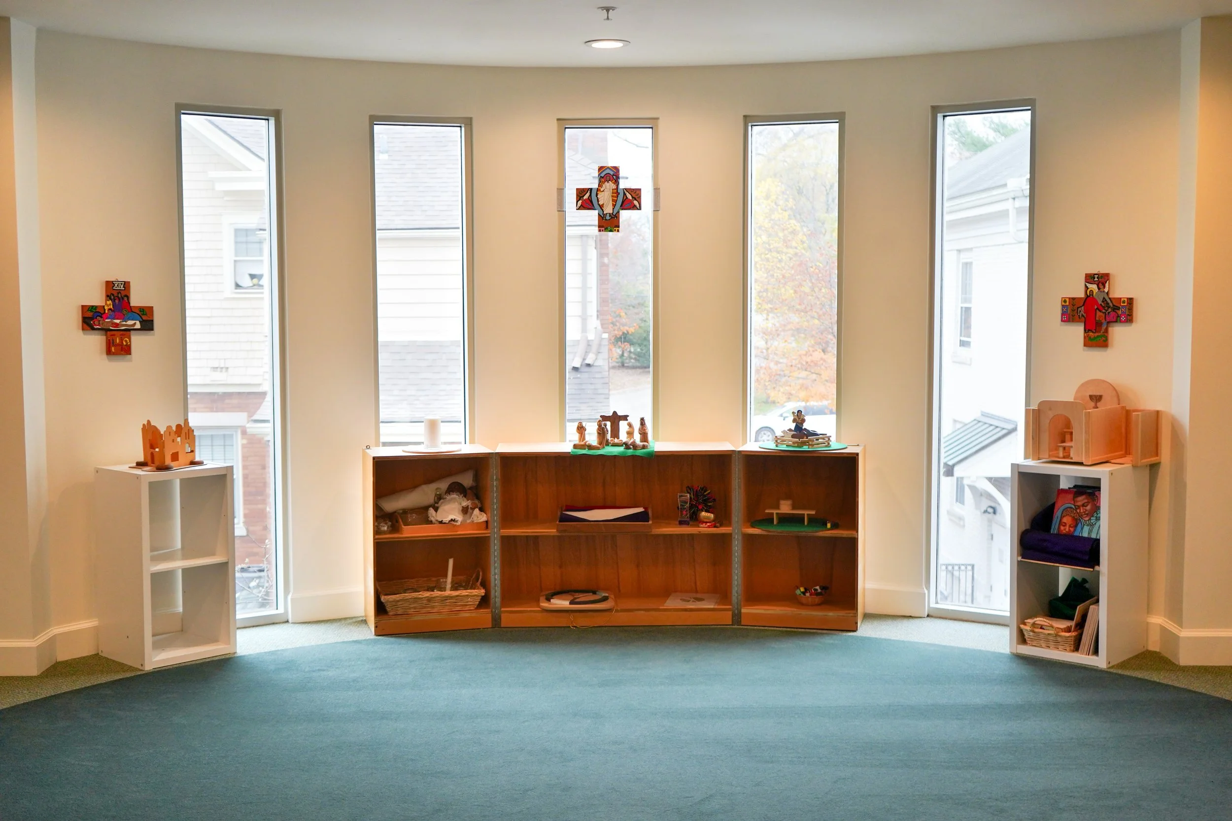 A room with a curved wall and five tall windows, decorated with colorful religious artwork and wooden shelves displaying Christian nativity figures and other religious items.