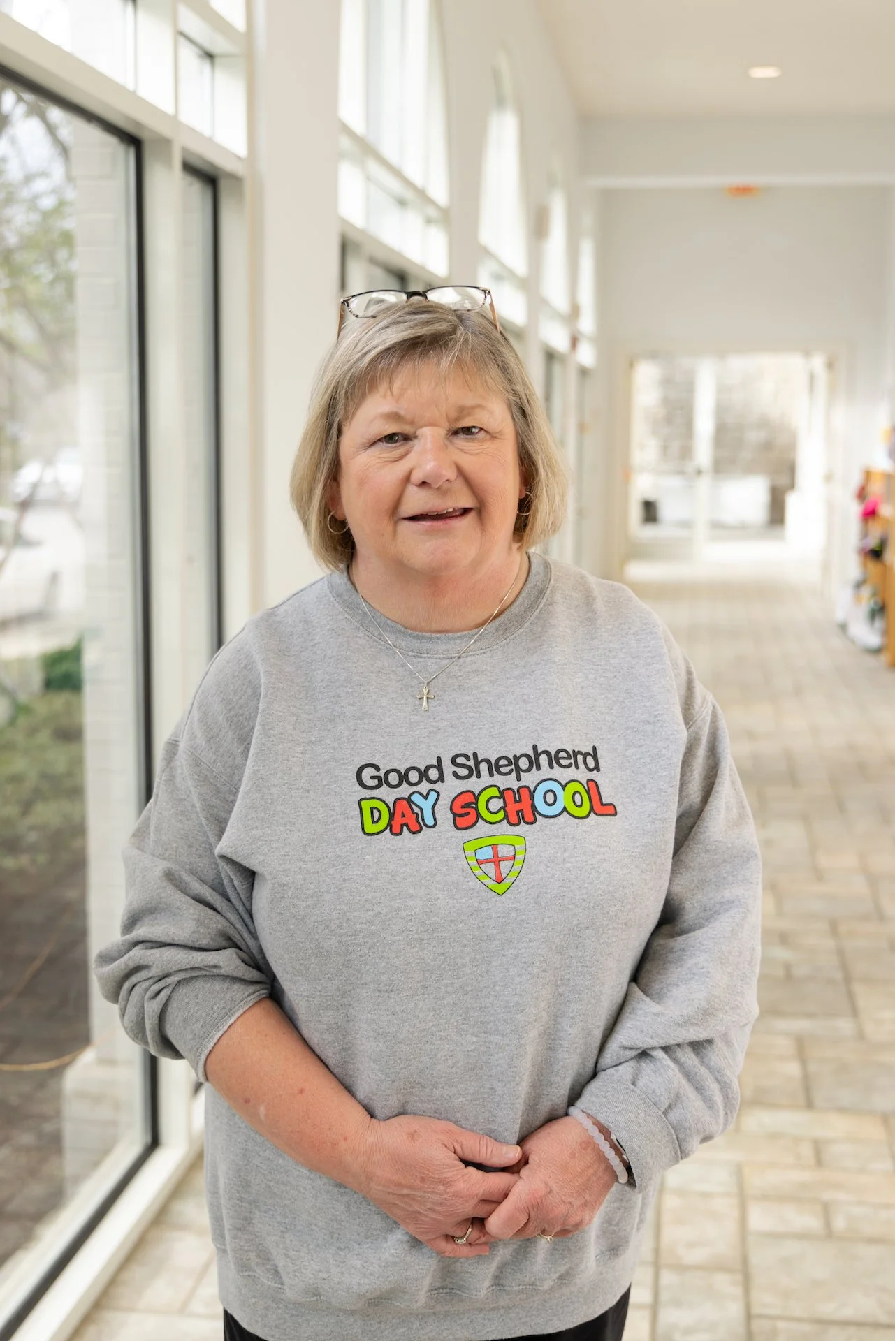 A smiling woman in a gray sweatshirt with 'Good Shepherd Day School' written on it standing indoors by large windows.