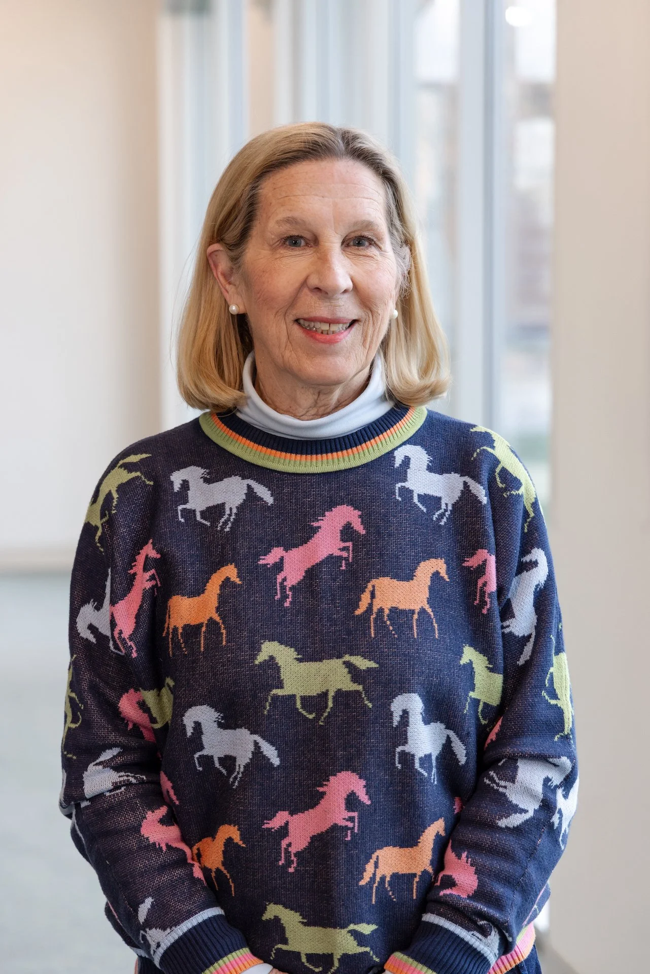 Older woman with shoulder-length blonde hair smiling, wearing a navy sweater with colorful horse patterns and a white turtleneck underneath, standing near a window.