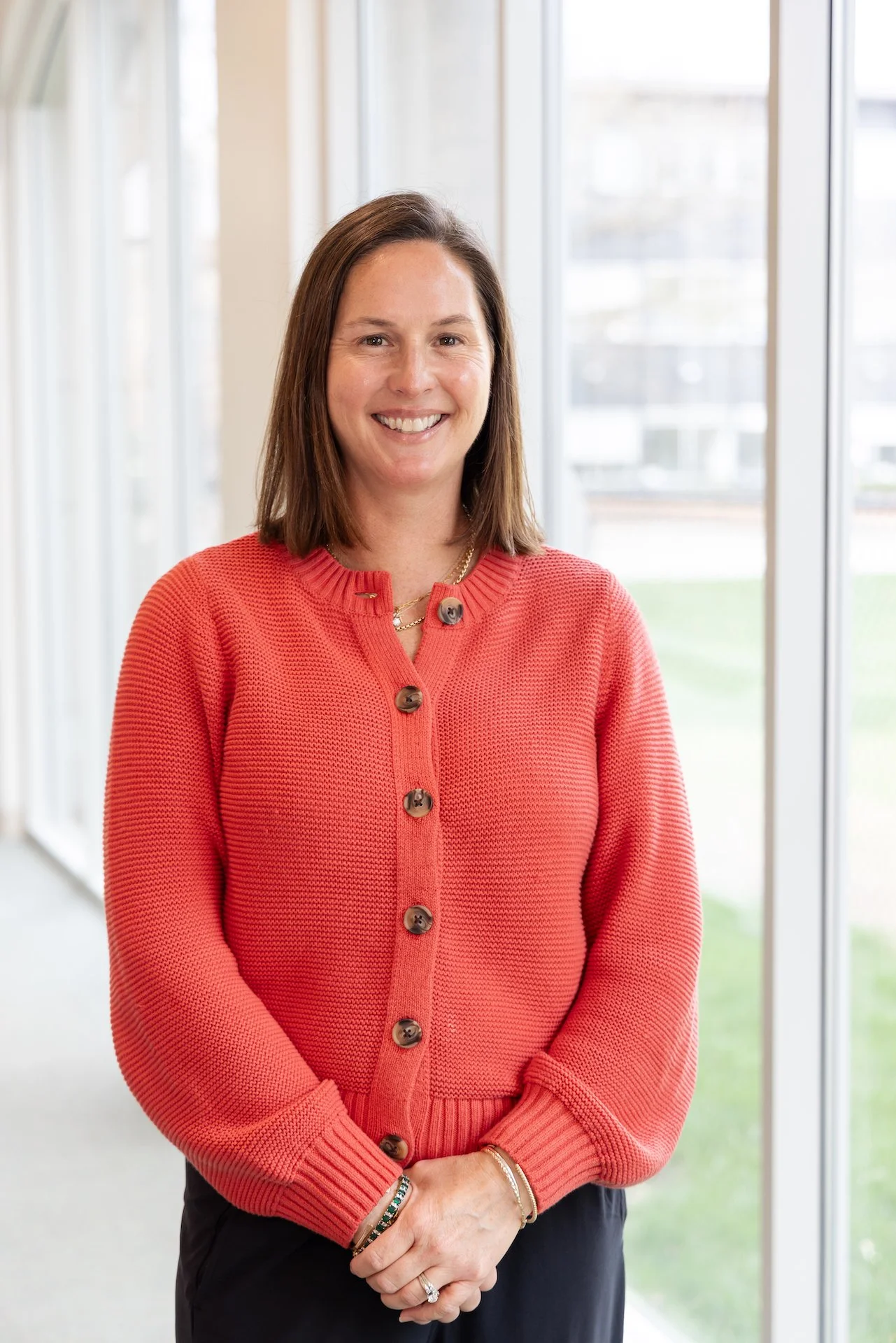 A woman smiling, standing indoors near large windows, wearing a red cardigan, gold jewelry, and dark pants.