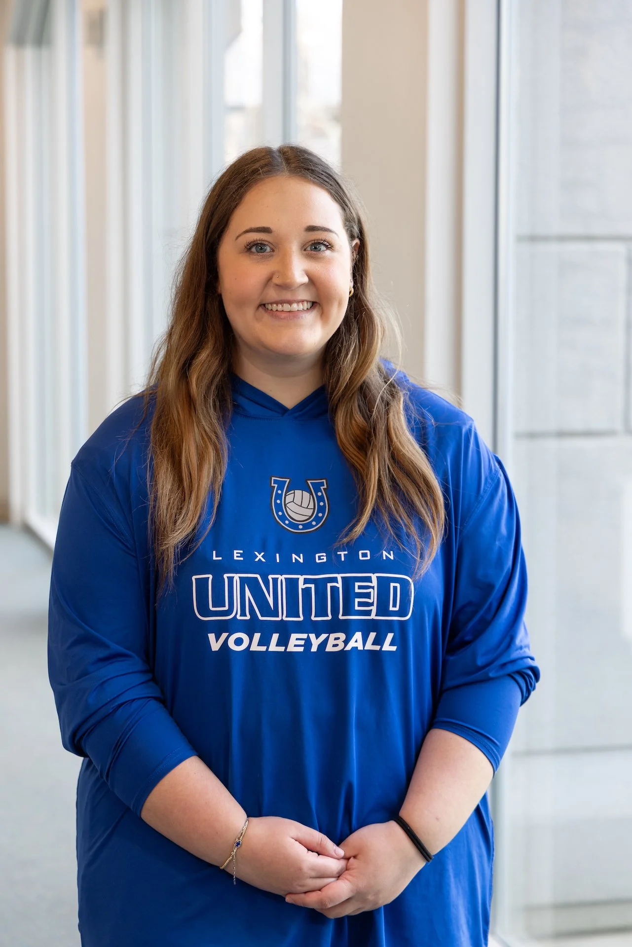 A young woman with long brown hair wearing a blue volleyball hoodie from Lexington United Volleyball, standing in an indoor setting with large windows.