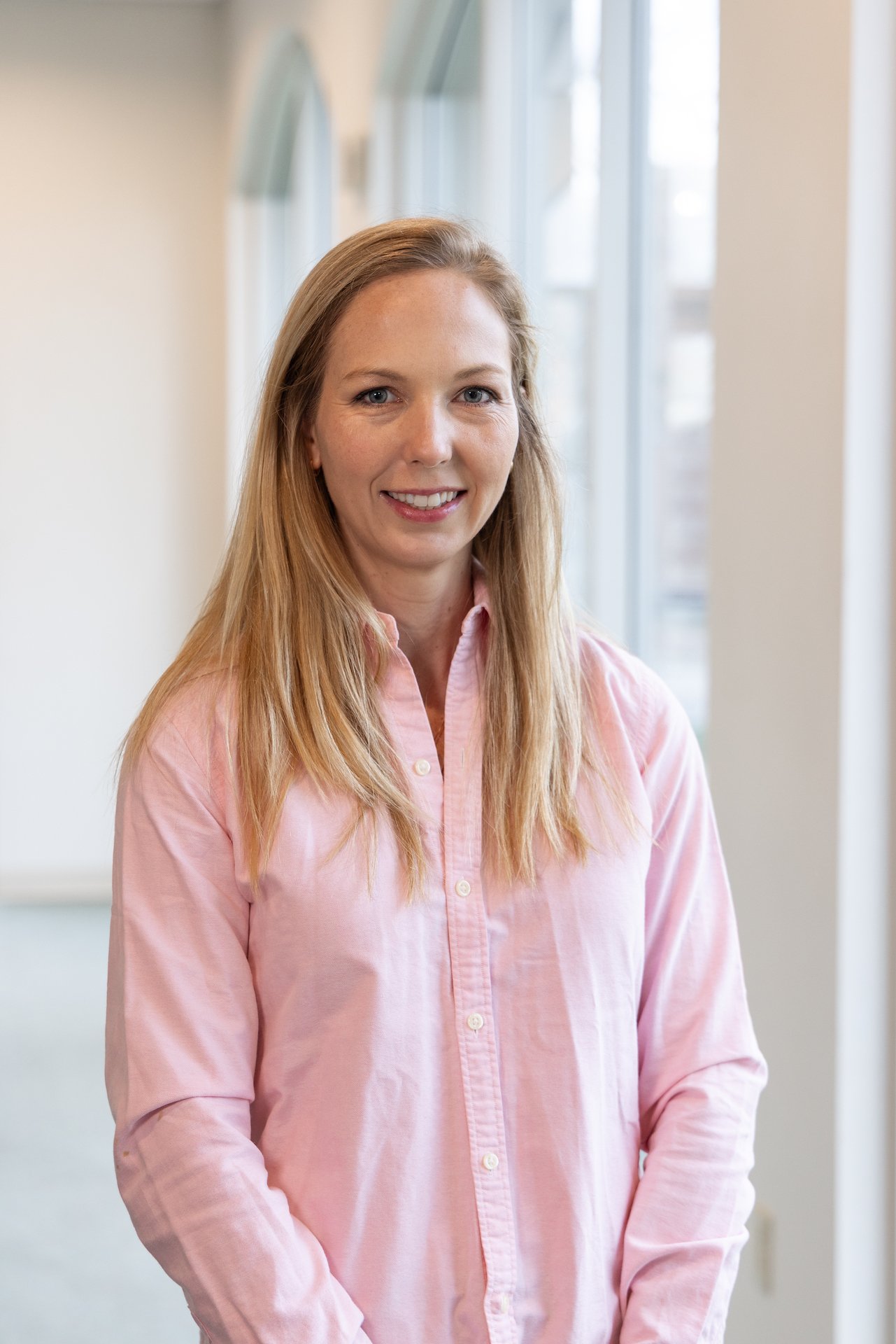 A woman with long blond hair wearing a light pink button-up shirt, smiling, standing indoors near large windows.