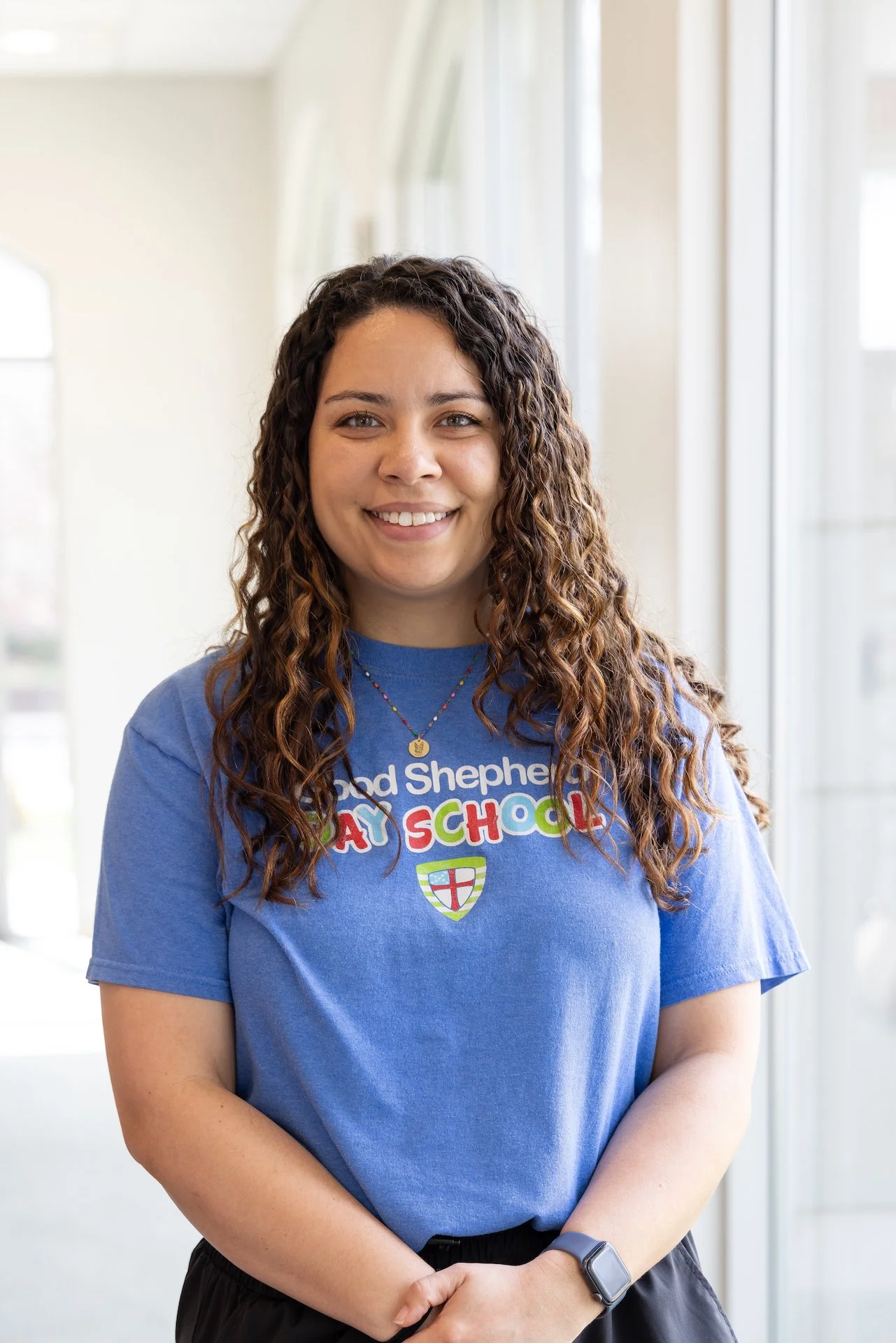 A woman with curly brown hair smiling, wearing a blue T-shirt that reads 'Good Shepherd Day School' with a colorful logo and a cross, standing indoors near large windows.