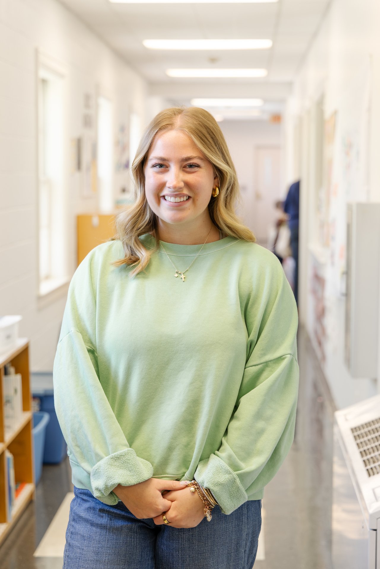 A young woman with blonde hair smiling, wearing a light green sweatshirt and blue jeans, standing in a hallway with white walls and windows.