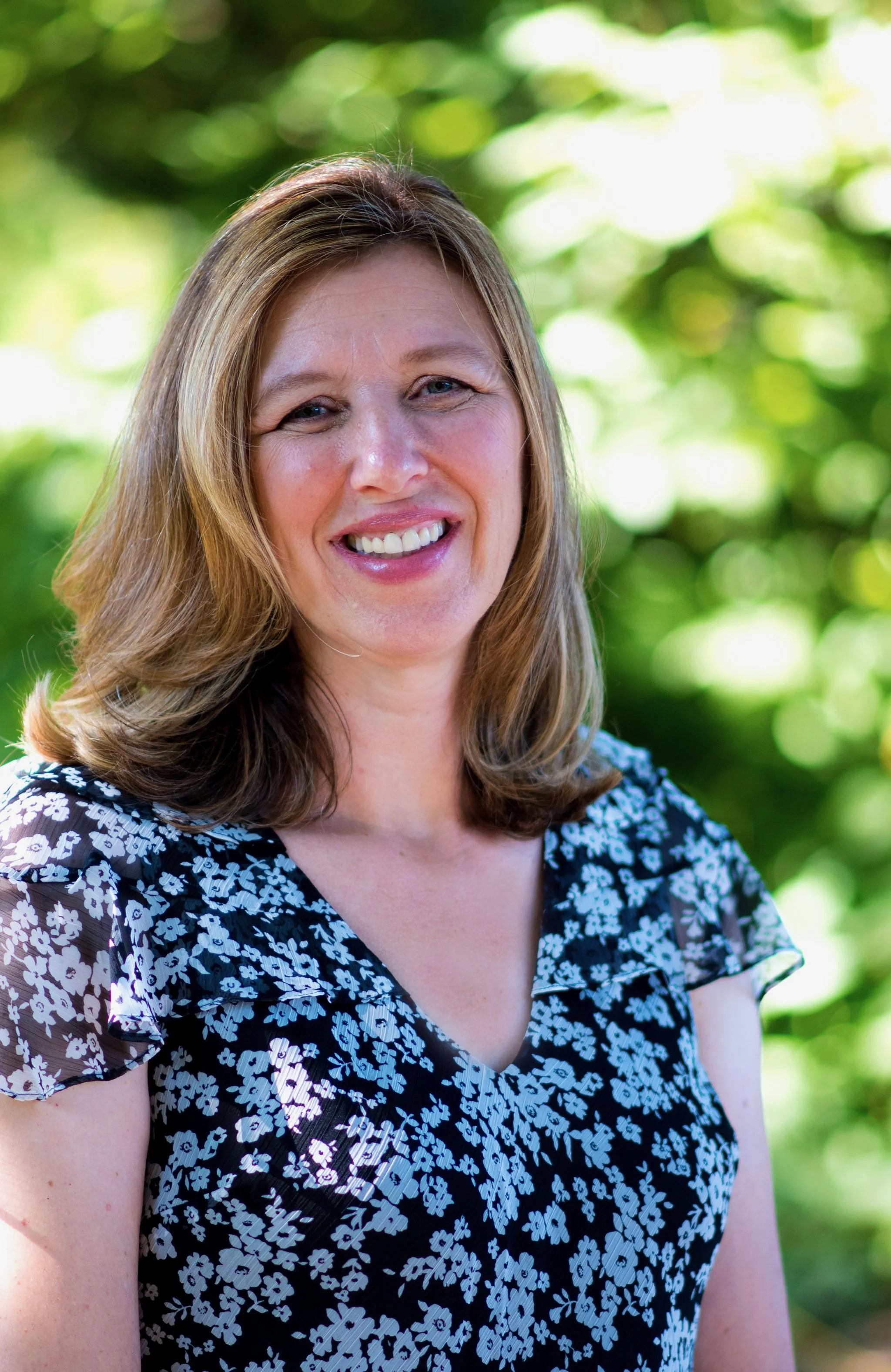 A woman with shoulder-length light brown hair smiling outdoors with a green, leafy background.