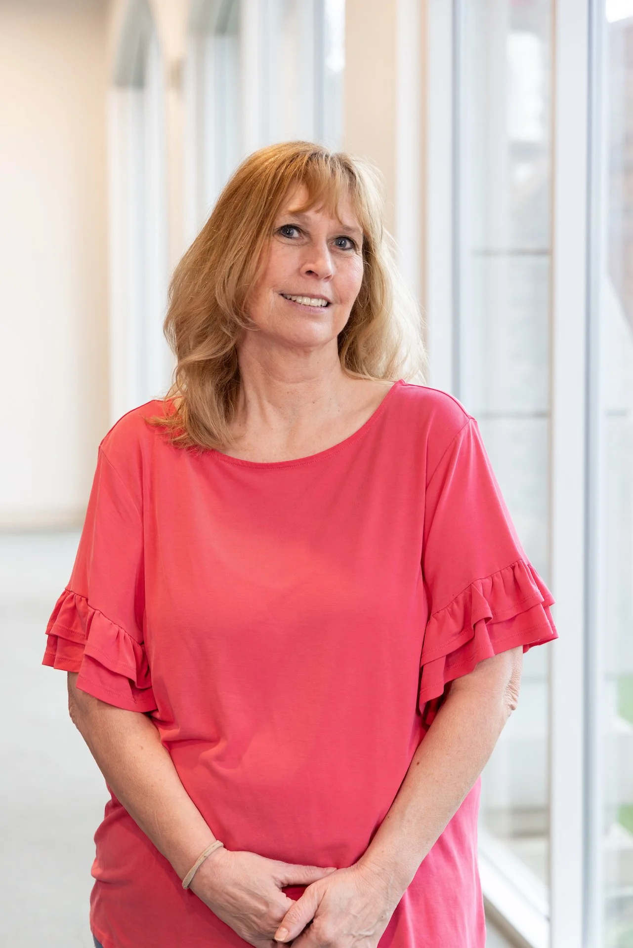 A woman with shoulder-length reddish-blonde hair wearing a pink top with ruffled sleeves, smiling and standing in front of large windows in a bright indoor setting.