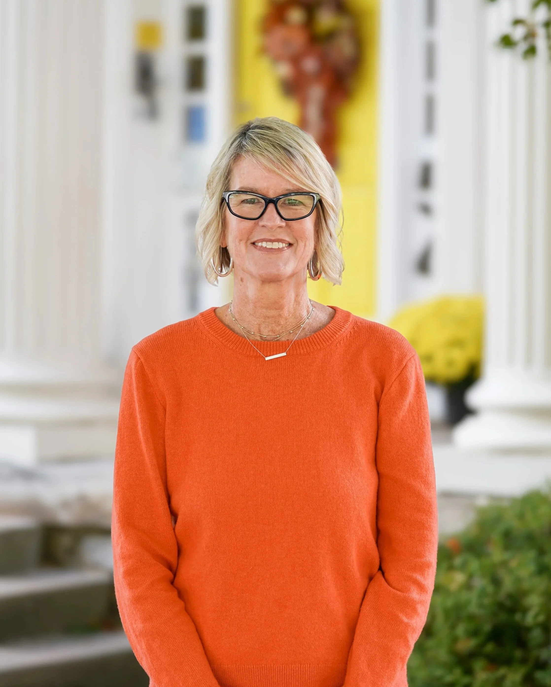 A woman with blond hair, glasses, earrings, and a necklace, smiling and wearing an orange sweater, standing indoors with colorful art and decor in the background.
