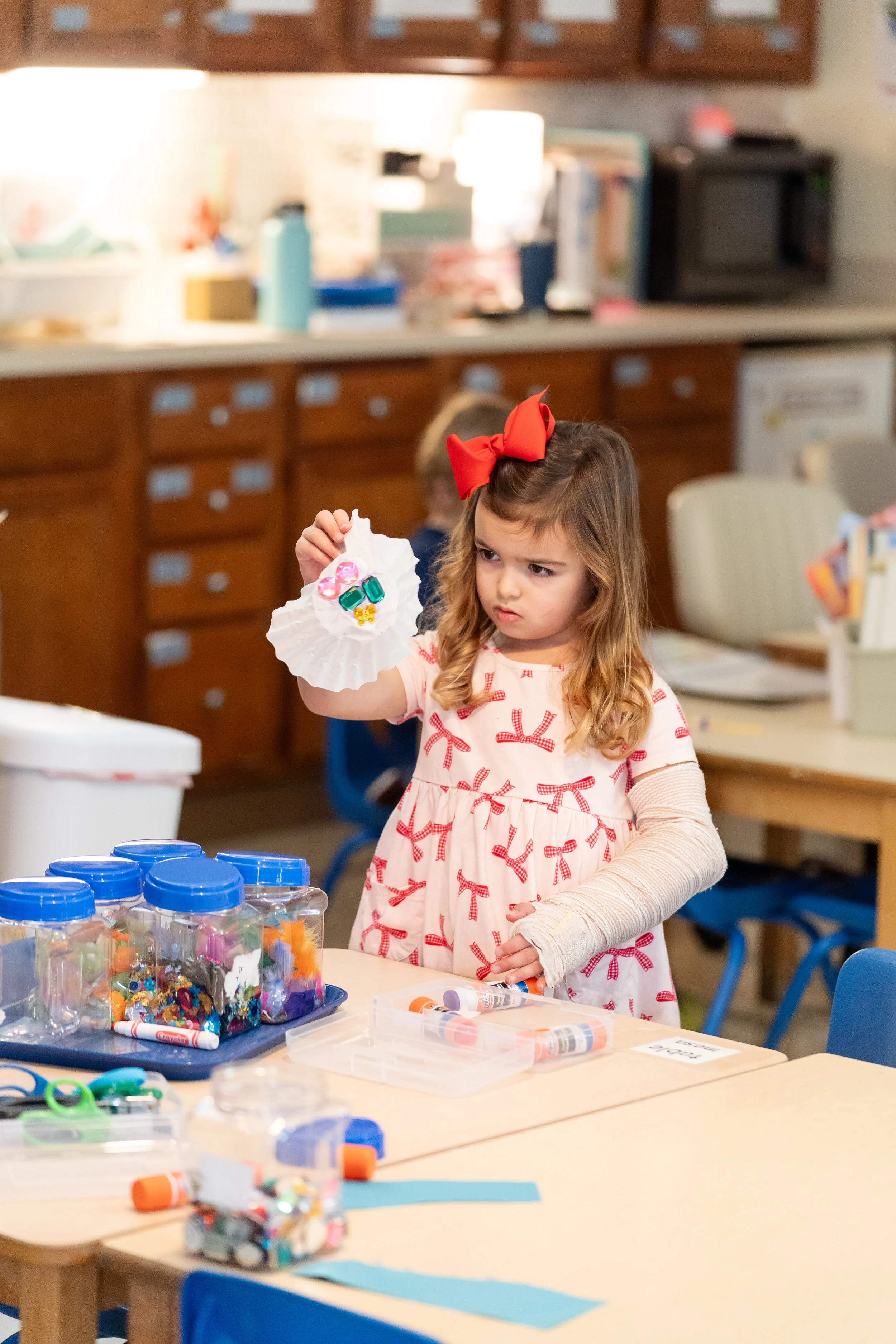 A young girl with long wavy hair, wearing a cream dress with red bows and a large red bow on her head, holding a paper fan decorated with colorful embellishments at a craft table in a classroom. The table has jars filled with craft supplies.