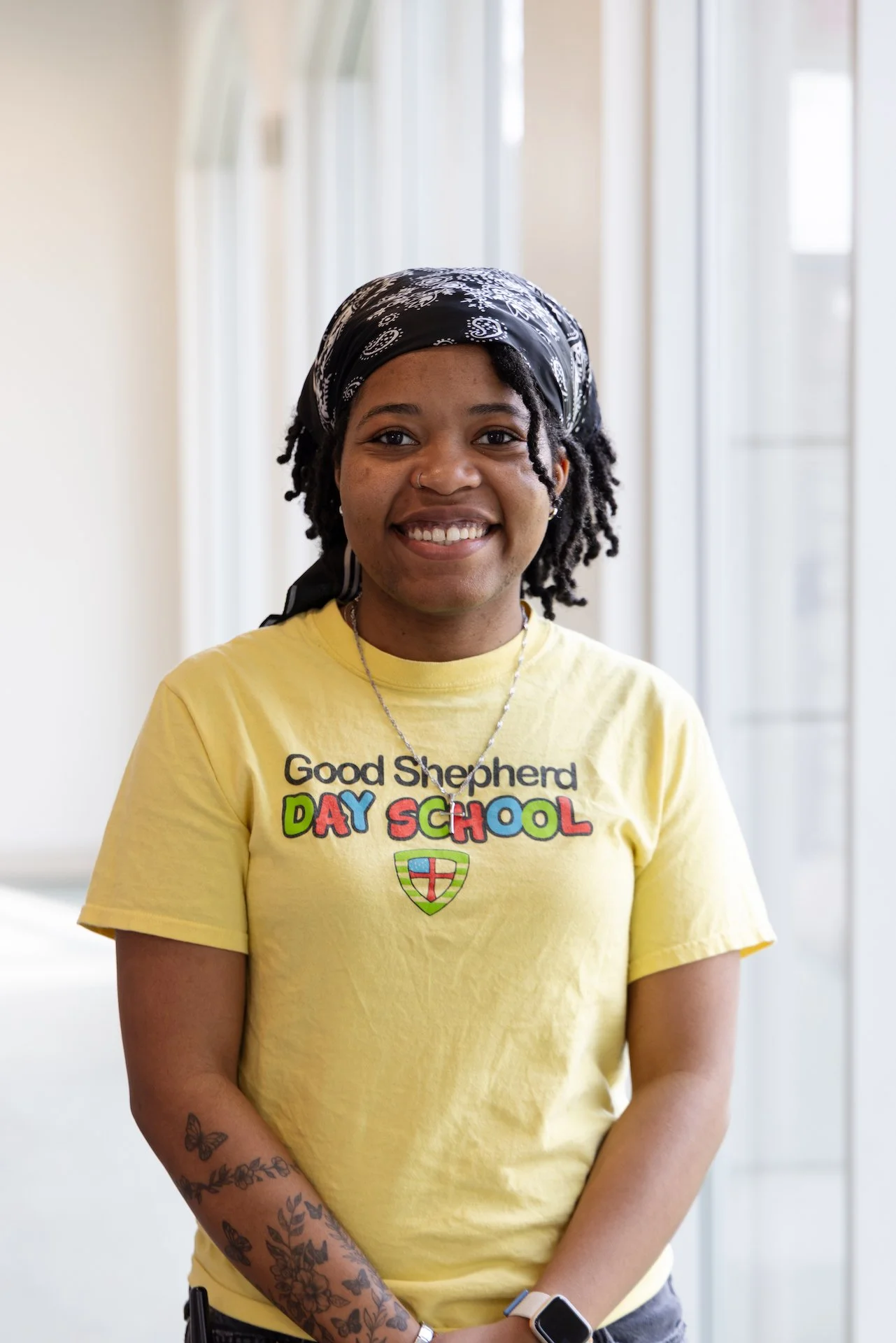 A young woman smiling, wearing a yellow T-shirt that reads 'Good Shepherd Day School', a black bandana, and a smartwatch, standing in front of a bright, sunlit background with windows and white framing.