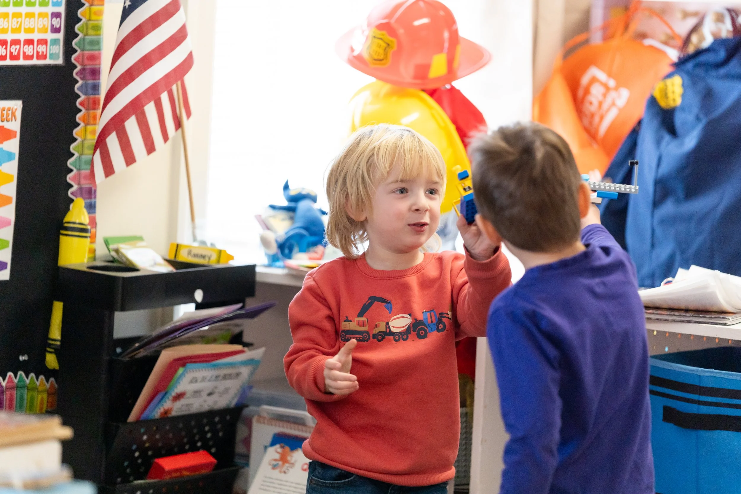 Two young boys in a classroom, one holding a toy boat and the other wearing a firefighter's helmet, surrounded by school supplies and colorful decorations.